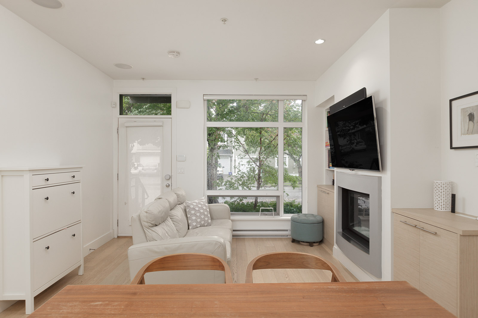 Modern living room with white walls, light wood flooring, a white sofa, wall-mounted TV above a fireplace, large windows, and minimal furniture.