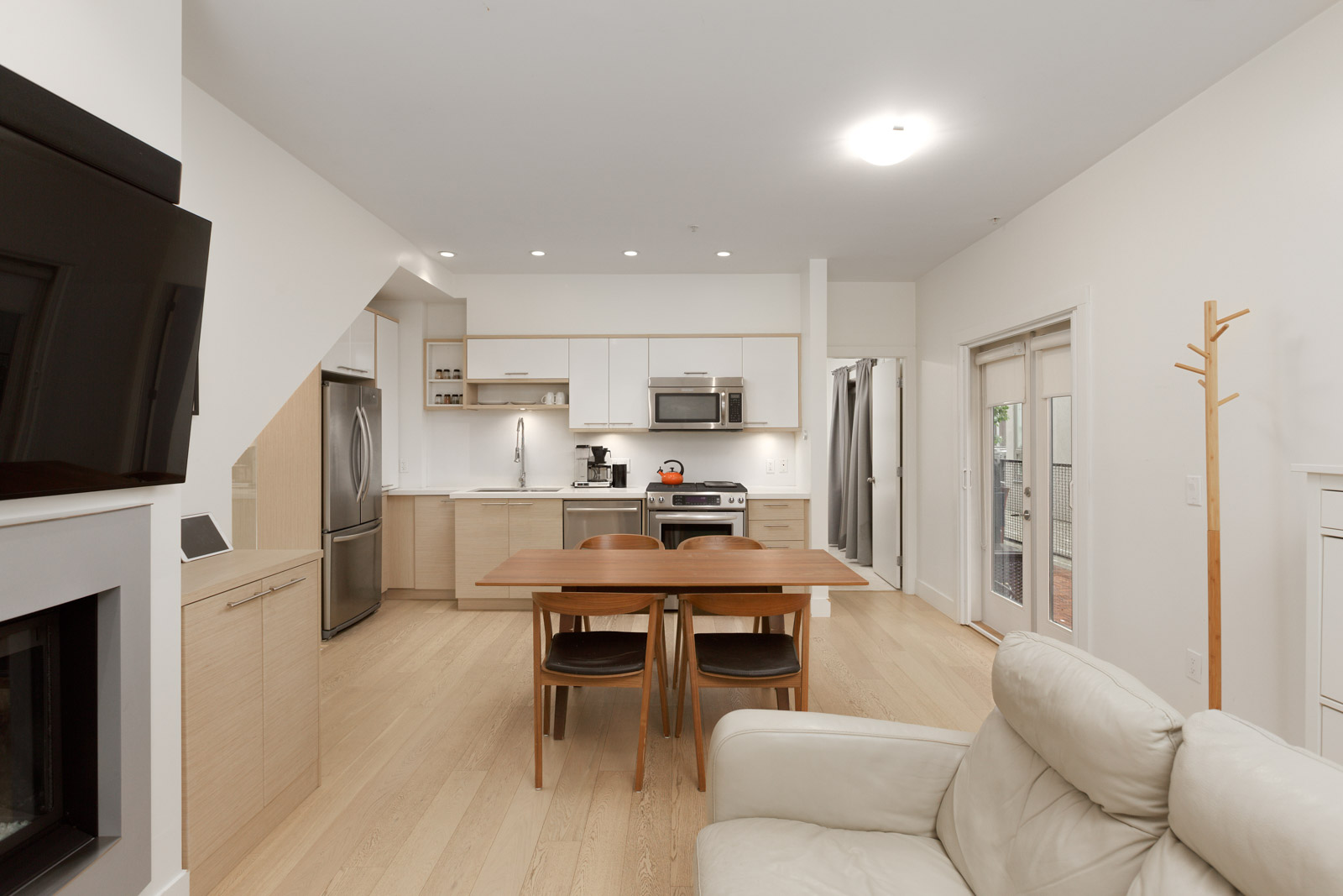 Modern open-plan kitchen and dining area with wooden floor, light cabinetry, stainless steel appliances, and a small dining table with four chairs. A white sofa is in the foreground.