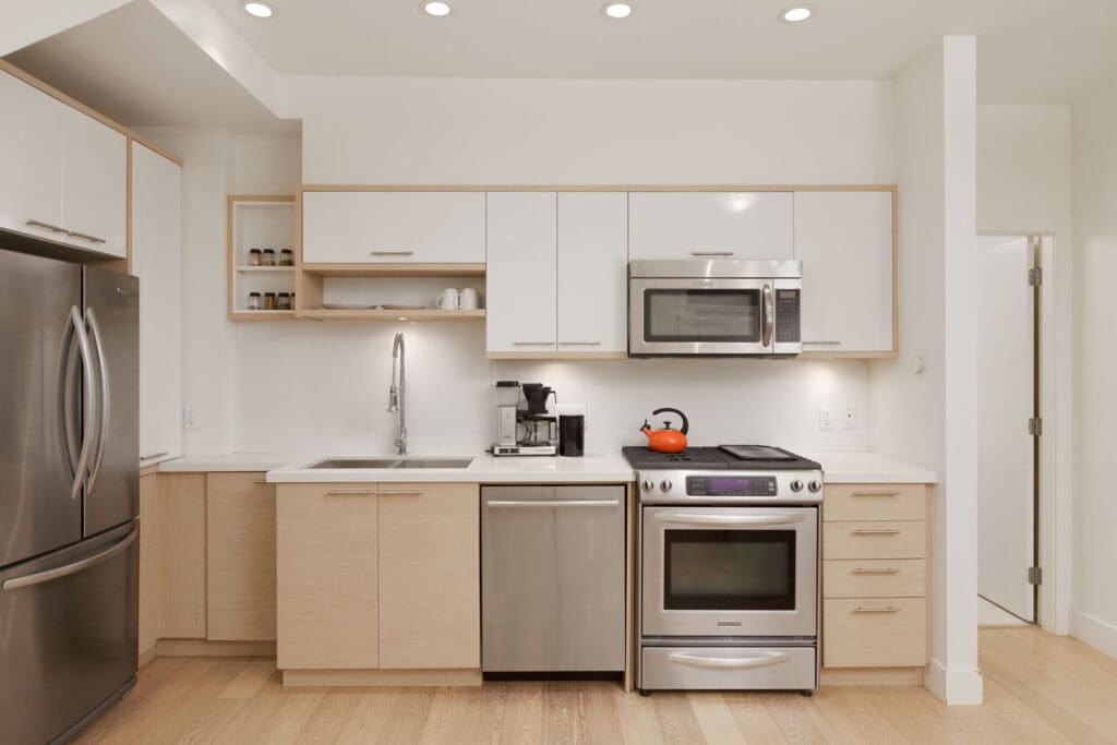 Modern kitchen with stainless steel appliances, white cabinets, light wood accents, and minimal decor. Counter holds a coffee maker and a red kettle sits on the stove.