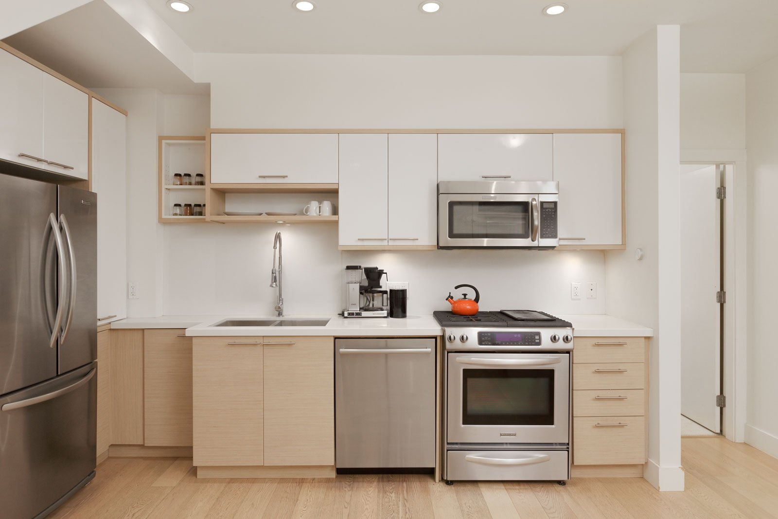 Modern kitchen with stainless steel appliances, white cabinets, light wood accents, and minimal decor. Counter holds a coffee maker and a red kettle sits on the stove.