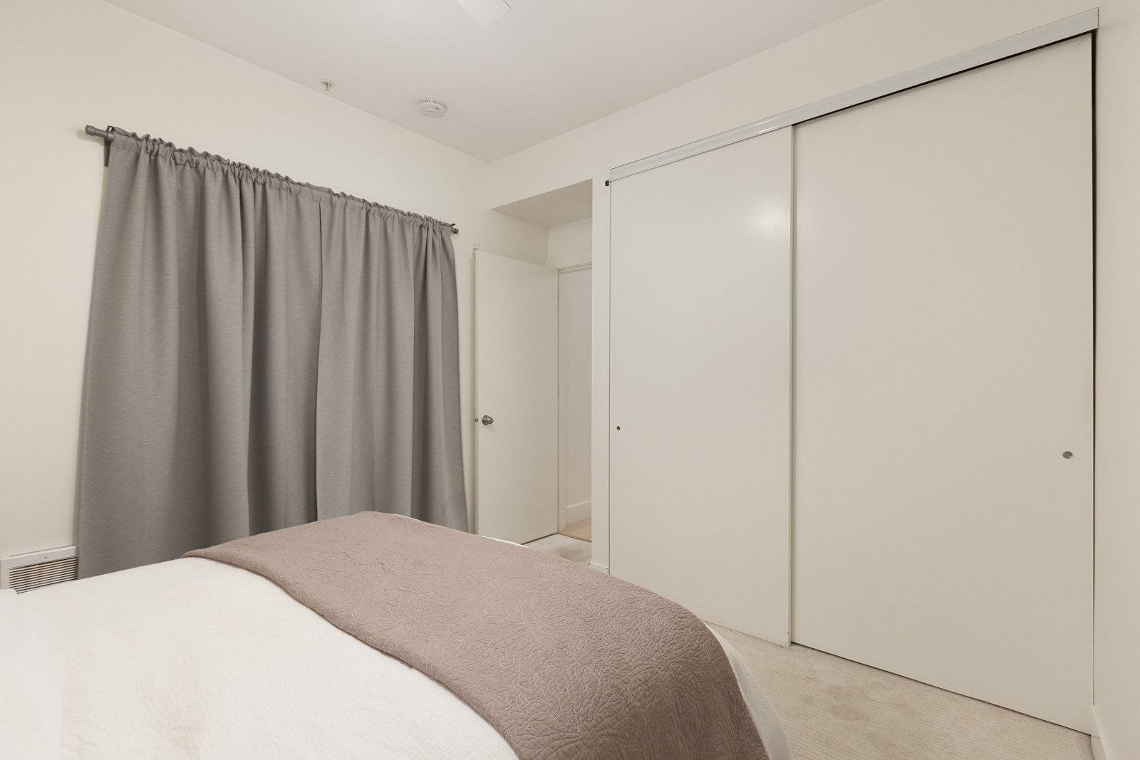 A bedroom with a bed, gray curtain covering a window, a white door, and sliding closet doors against white walls.