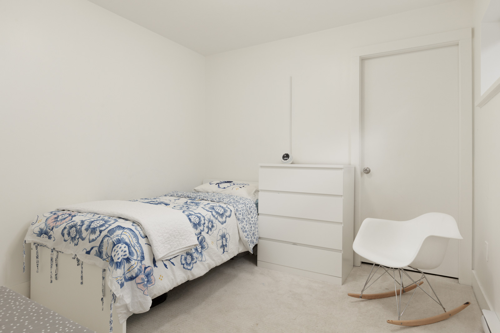 A small white bedroom with a single bed, blue floral bedding, a white dresser, and a white modern rocking chair.