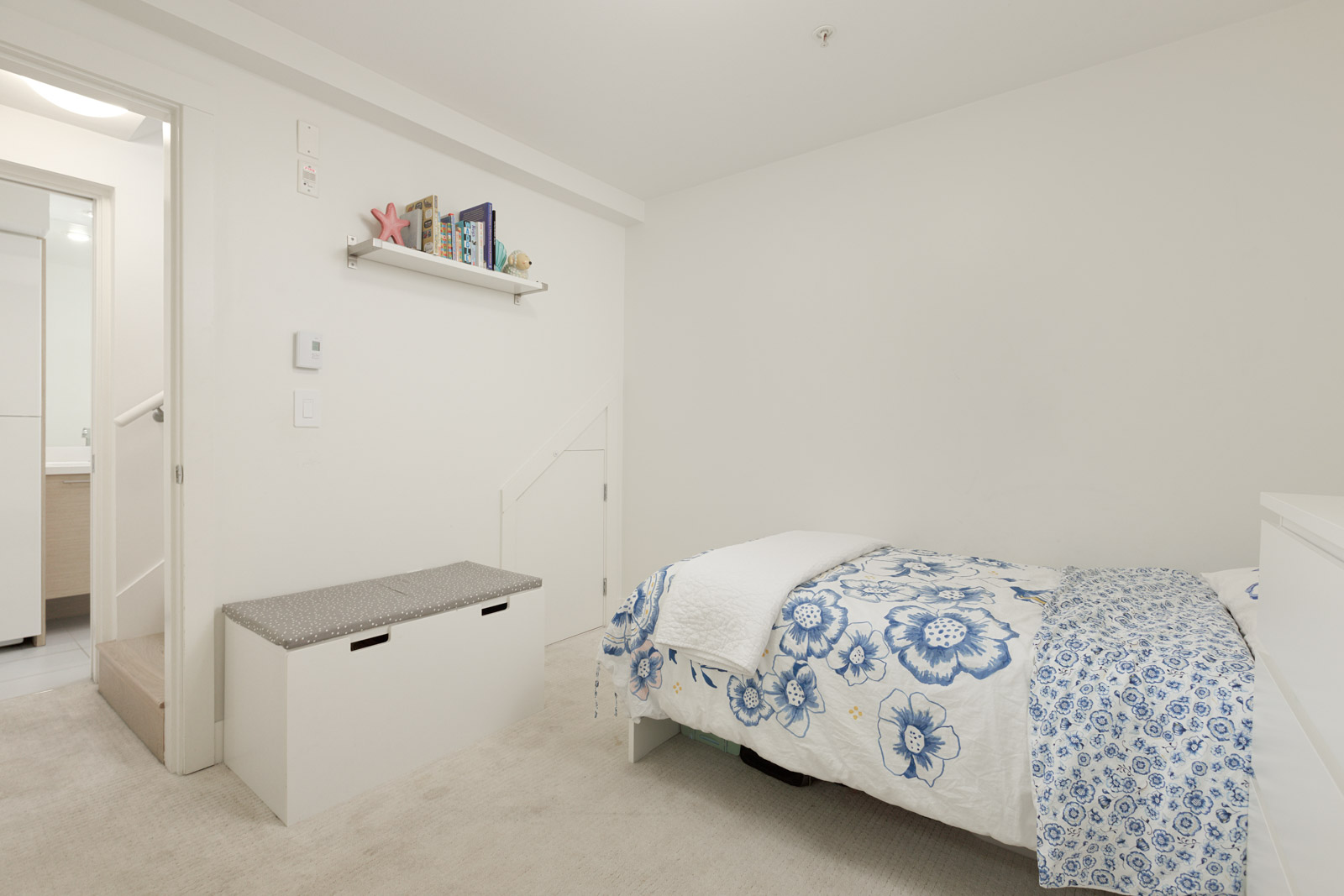 A small, white bedroom with a bed featuring blue and white floral bedding, a bench with storage, a wall shelf, and a door leading to a bathroom.