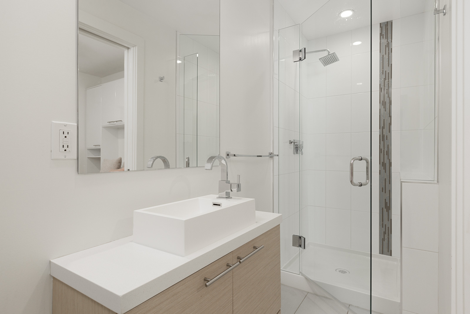 Modern bathroom with a rectangular sink on a wood vanity, large wall mirror, and a glass-enclosed shower with white tiles and a vertical accent strip.