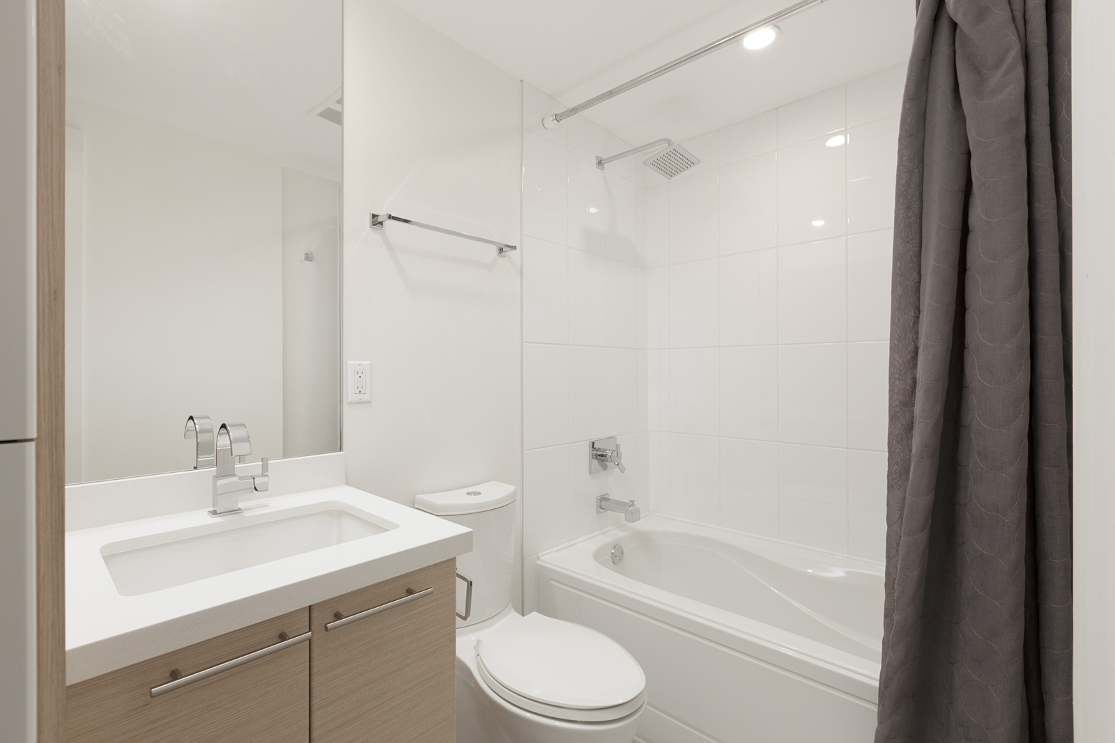 Modern bathroom with a white sink, light wood vanity, toilet, and bathtub-shower combination with white tiles and a gray shower curtain. Bright overhead lighting.