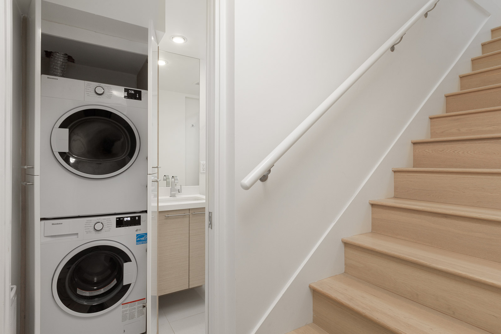 A stacked washer and dryer are in a small laundry closet next to a bathroom and a light wood staircase.