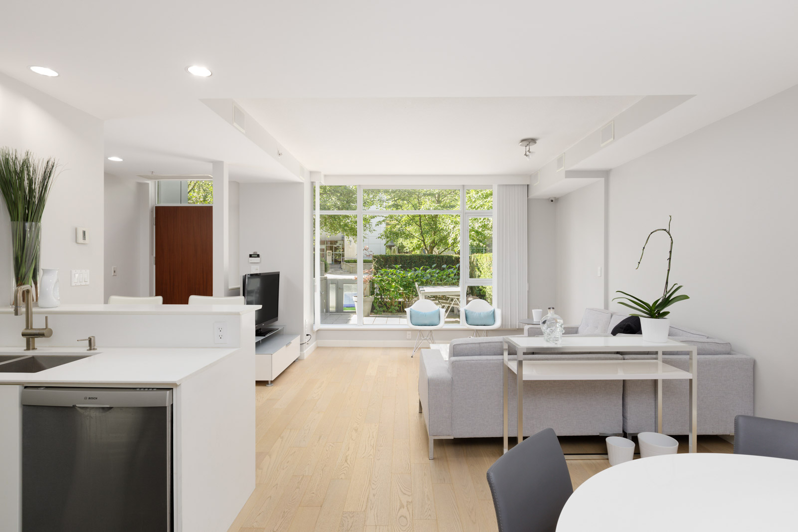 Modern, open-concept living room and kitchen with light wood floors, white walls, large windows, grey sofa, and greenery visible outside.