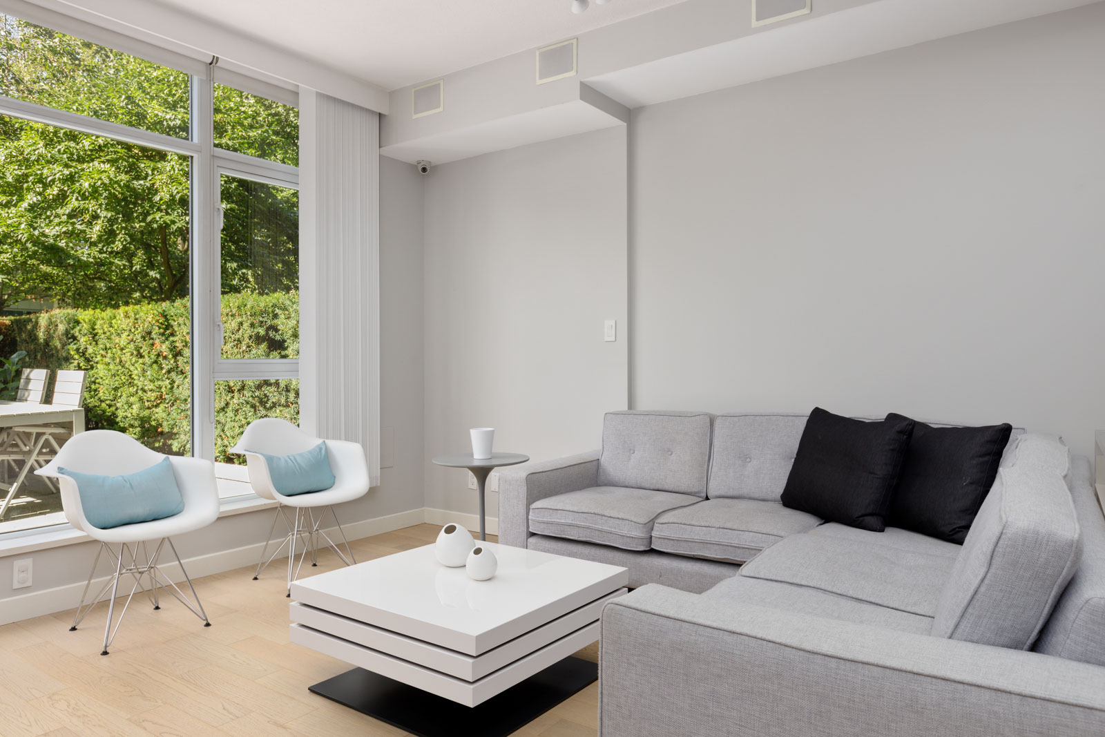 Modern living room with a gray sectional sofa, black and light blue accent pillows, two white chairs, a white coffee table, and large window overlooking greenery.