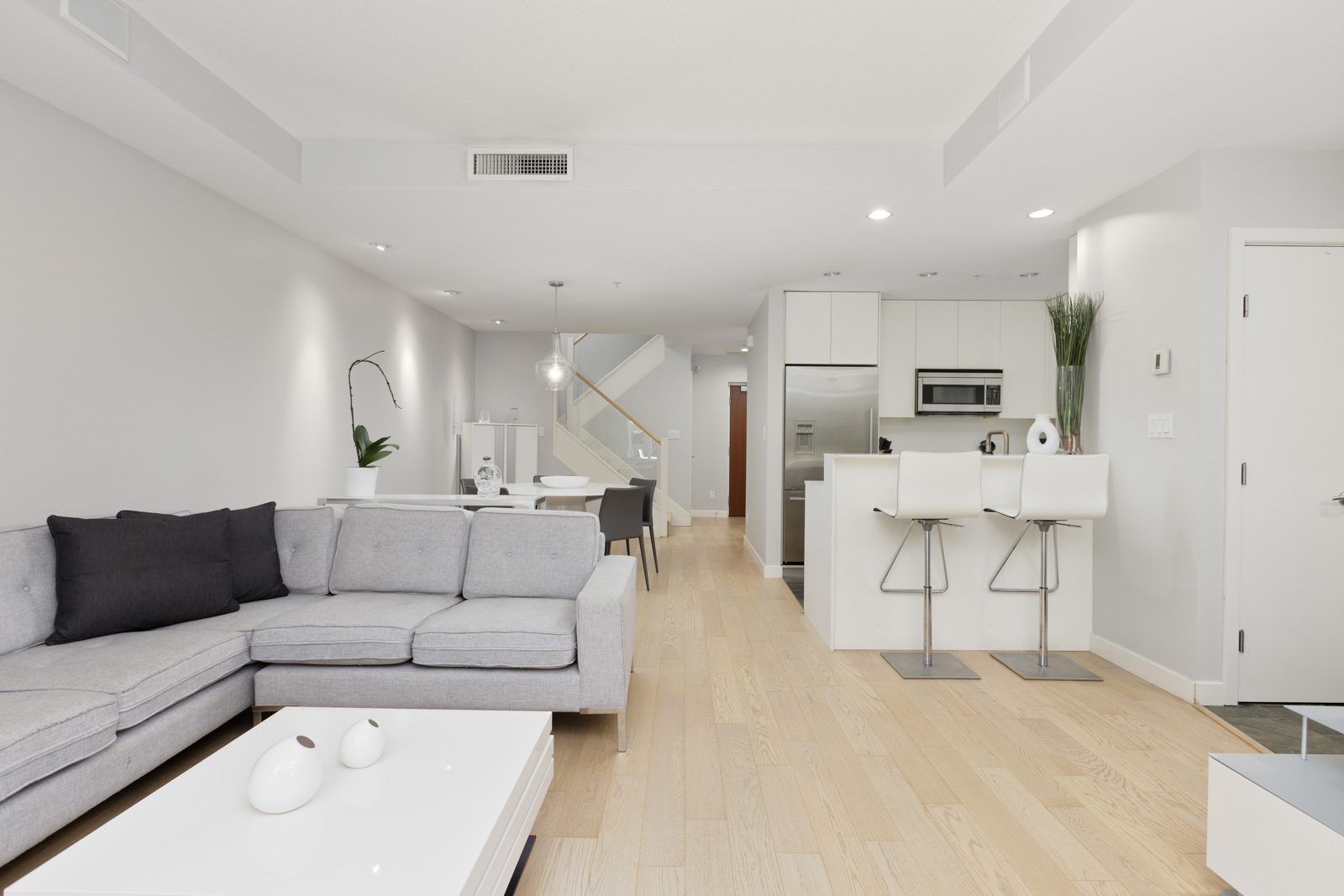 Modern open-plan living area with light wood flooring, grey sectional sofa, white kitchen, breakfast bar with two stools, and dining area in the background.