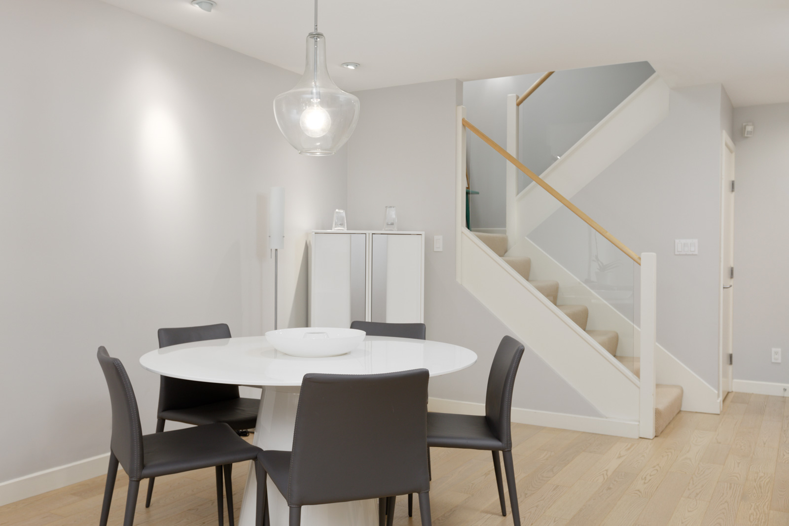 A modern dining area with a round white table, four black chairs, a hanging light fixture, and a staircase in the background.