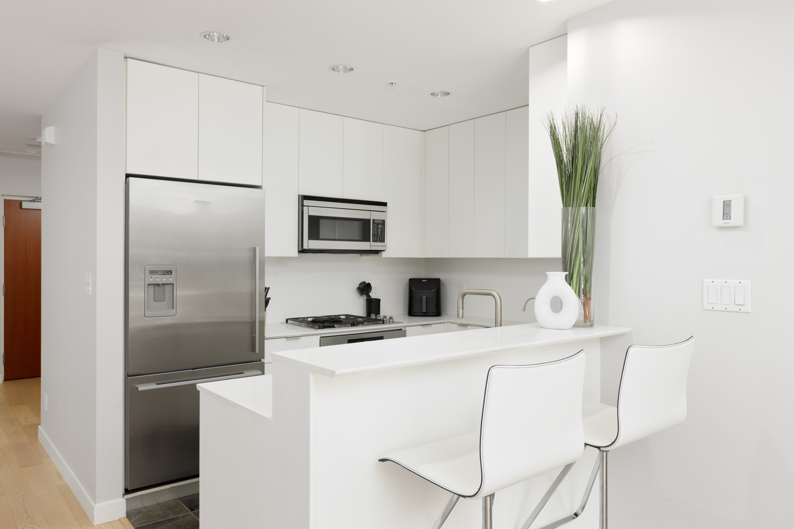 Modern white kitchen with stainless steel appliances, two white bar stools at a breakfast counter, and a tall vase with decorative grass on the counter.