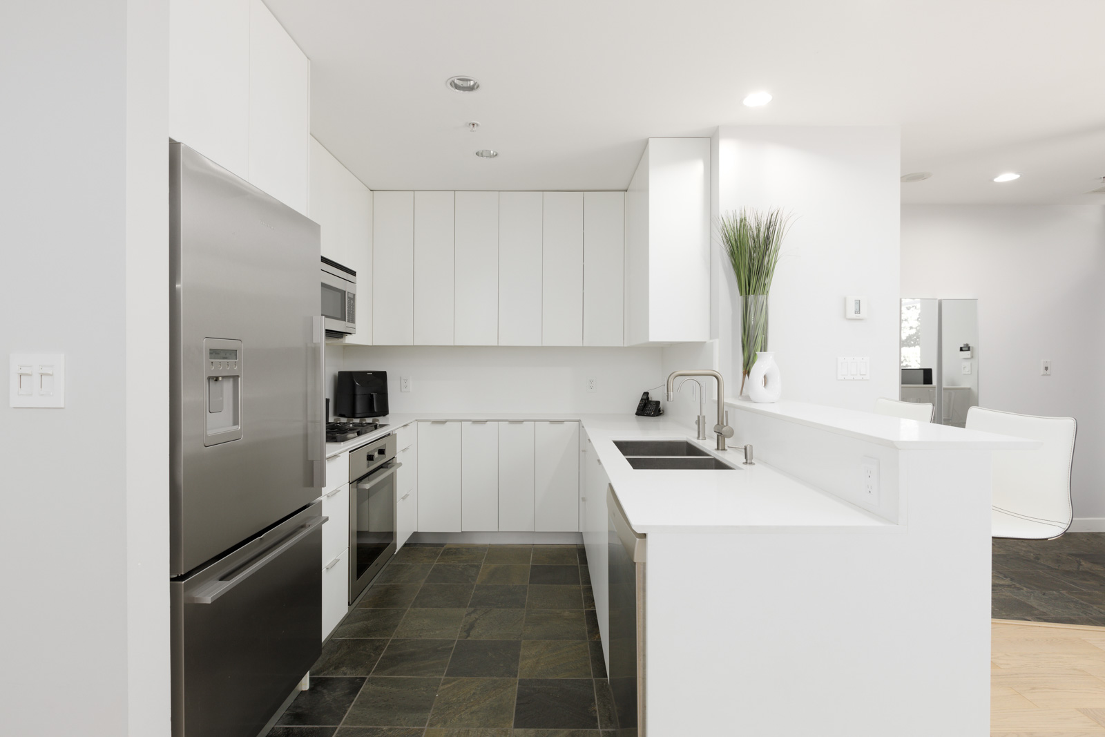 Modern kitchen with white cabinets, stainless steel appliances, slate tile flooring, a black sink, and minimalist decor including a tall vase with green grass.
