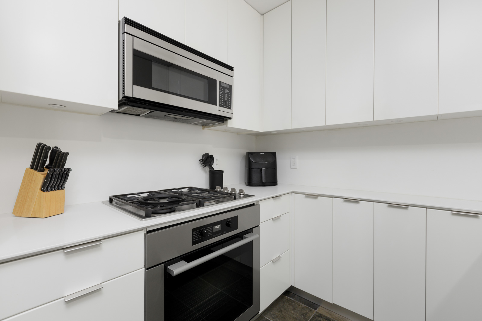 Modern kitchen with white cabinets, a built-in oven, a gas stove, a microwave, a knife block, and a coffee maker on a white countertop.