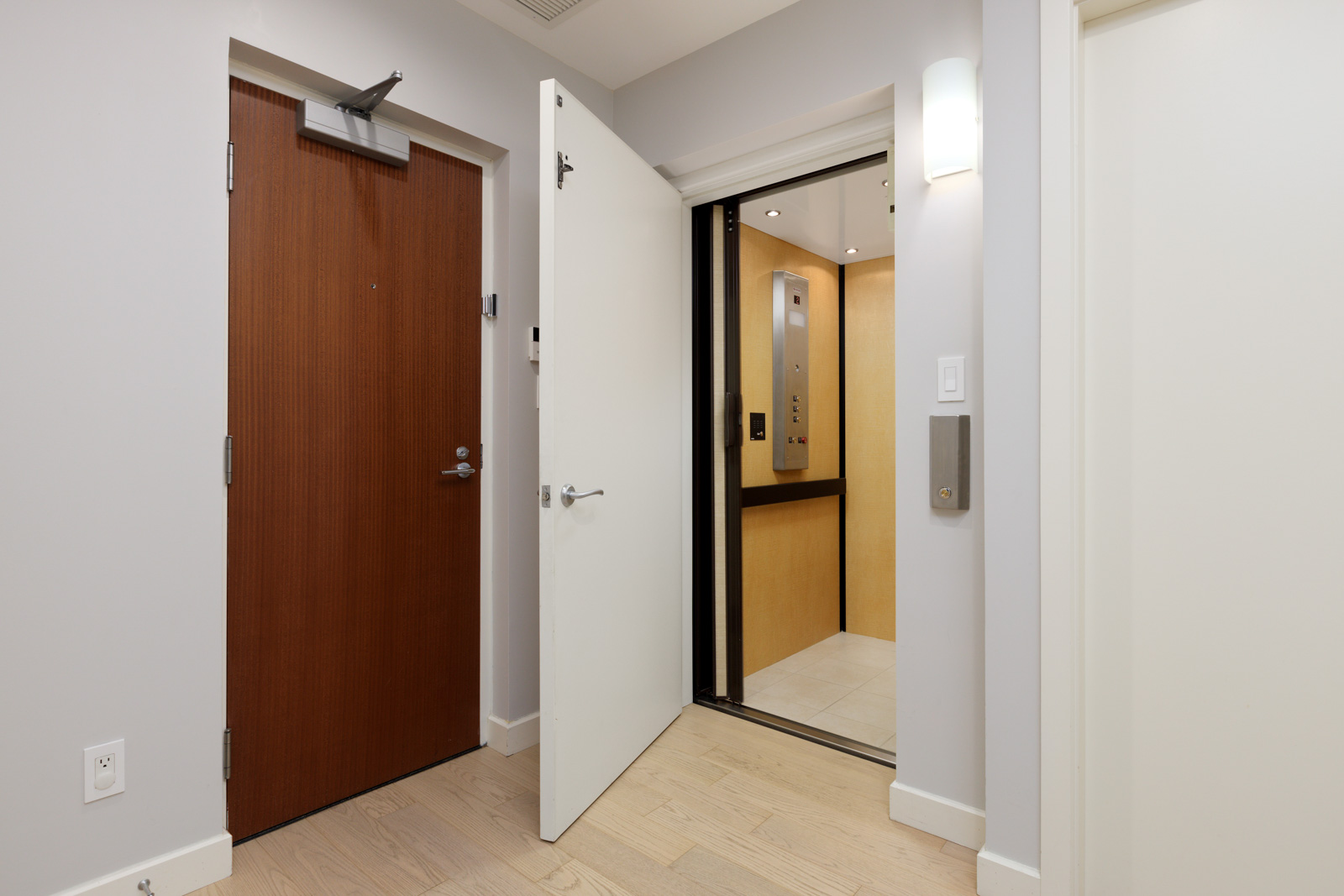 A modern hallway with a wooden front door and an open white door leading to an elevator with beige interior walls.