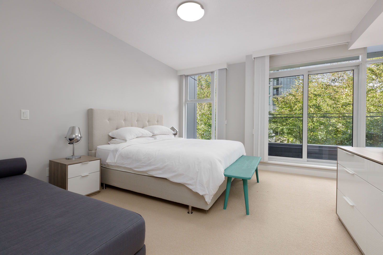 Modern bedroom with a large bed, nightstand, green bench, sofa, dresser, and floor-to-ceiling windows overlooking trees.