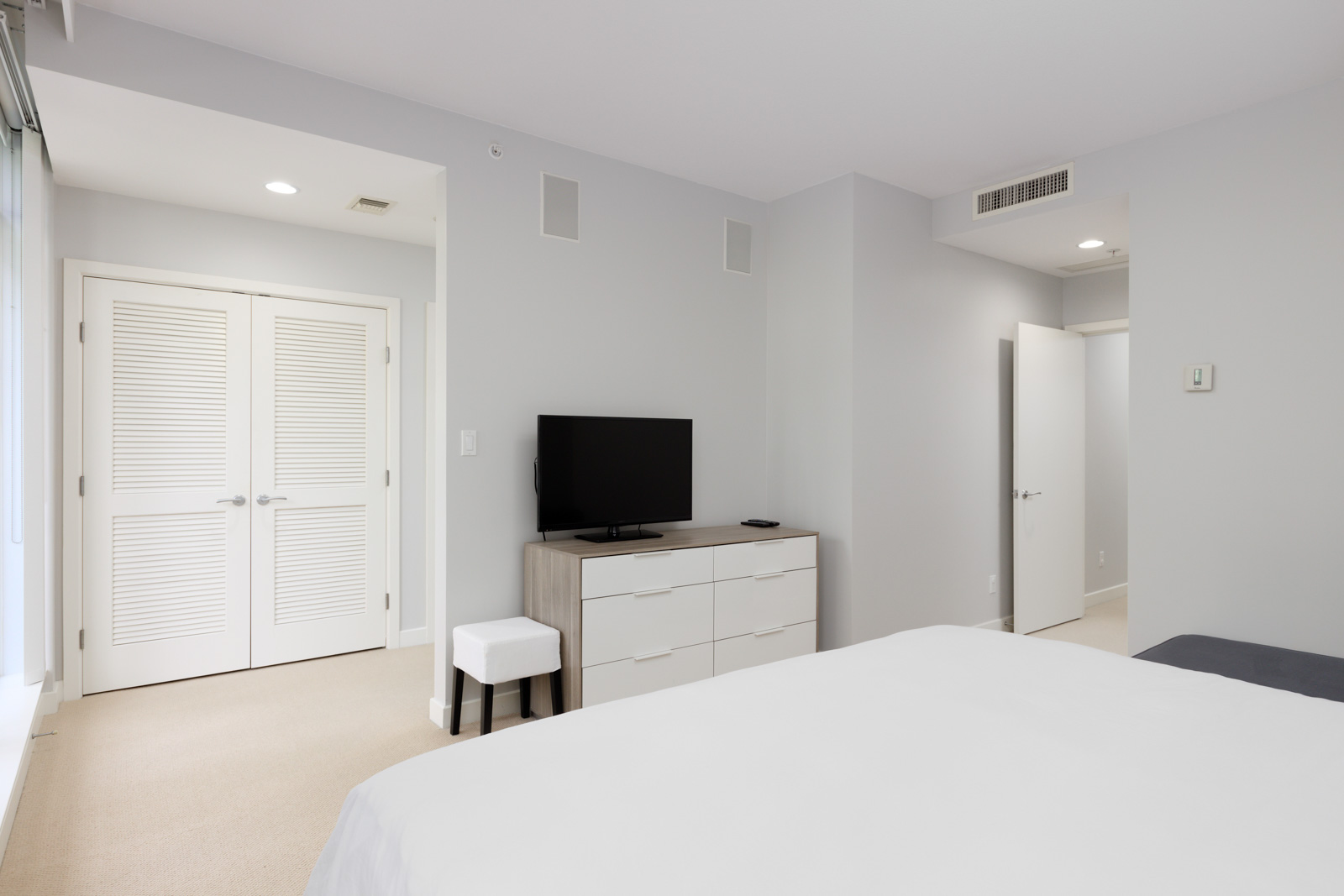 A modern bedroom with light walls, a white bed, dresser with TV, stool, double closet doors, and an open door leading to a hallway.