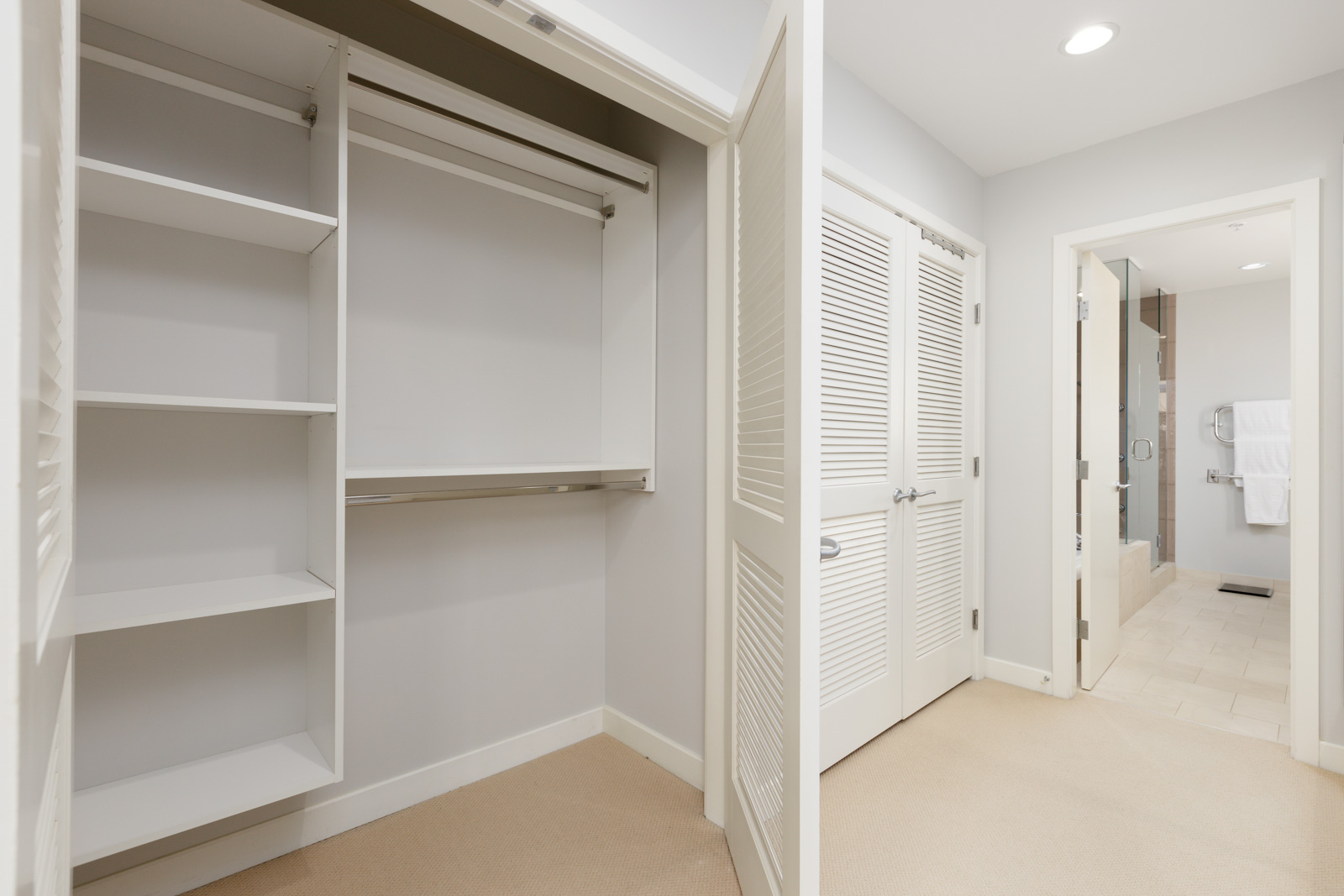 Empty walk-in closet with open louvered doors, white shelves, and hanging rods, adjacent to a hallway leading to a bathroom with tiled floor.