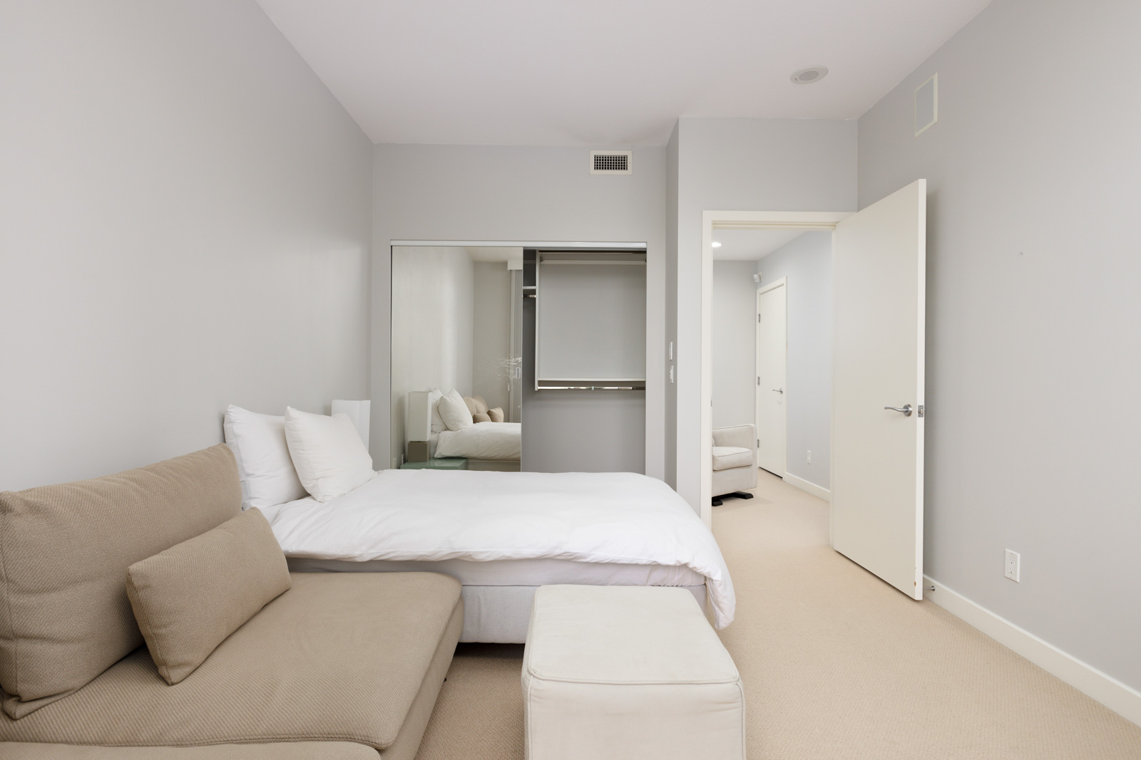 A minimalist bedroom with a bed, beige sofa, white ottoman, mirrored closet doors, and neutral walls and carpet. An open door leads to a hallway with additional rooms visible.