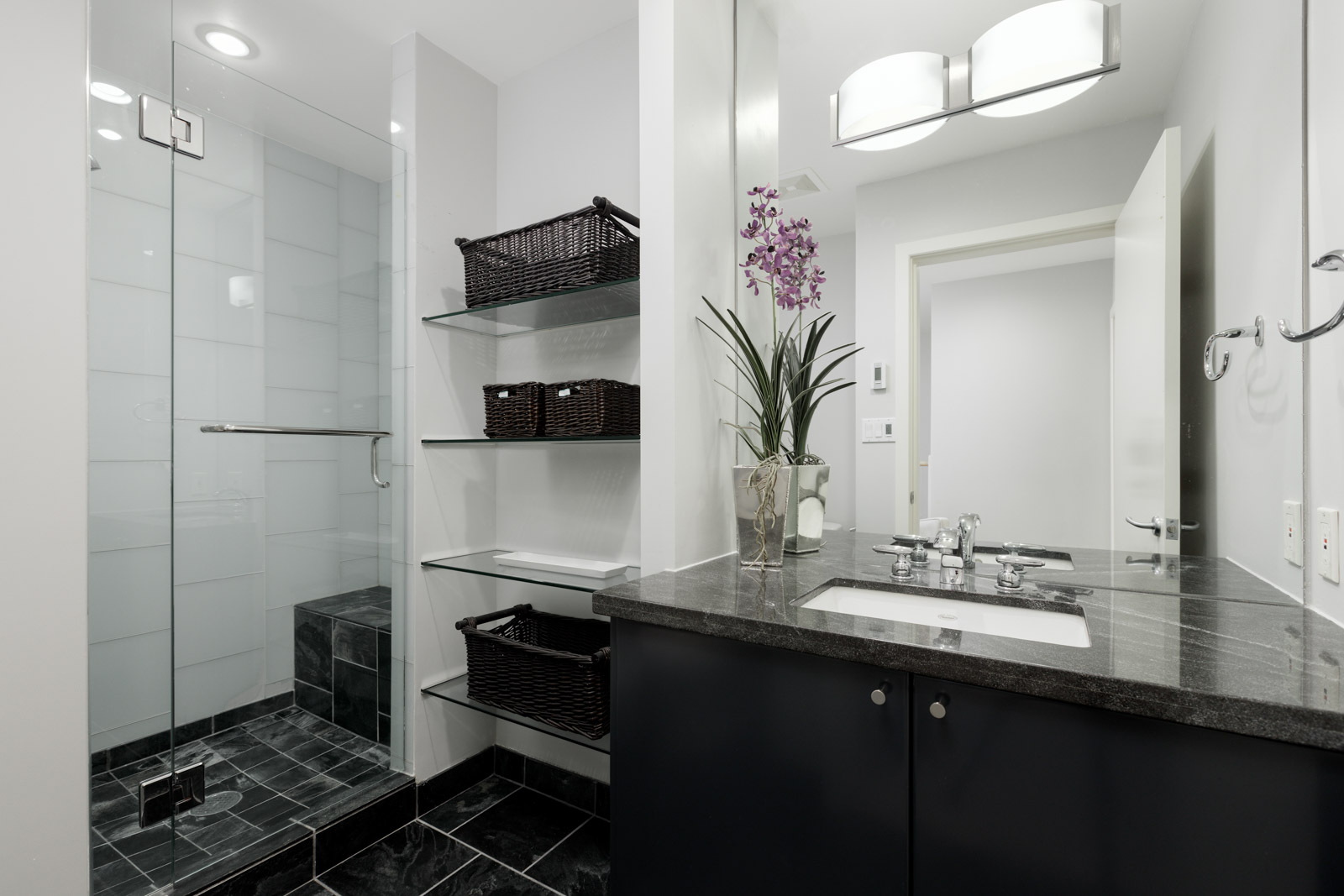 Modern bathroom with a walk-in glass shower, black countertop sink, glass shelves with dark baskets, and a vase with purple flowers by the mirror.