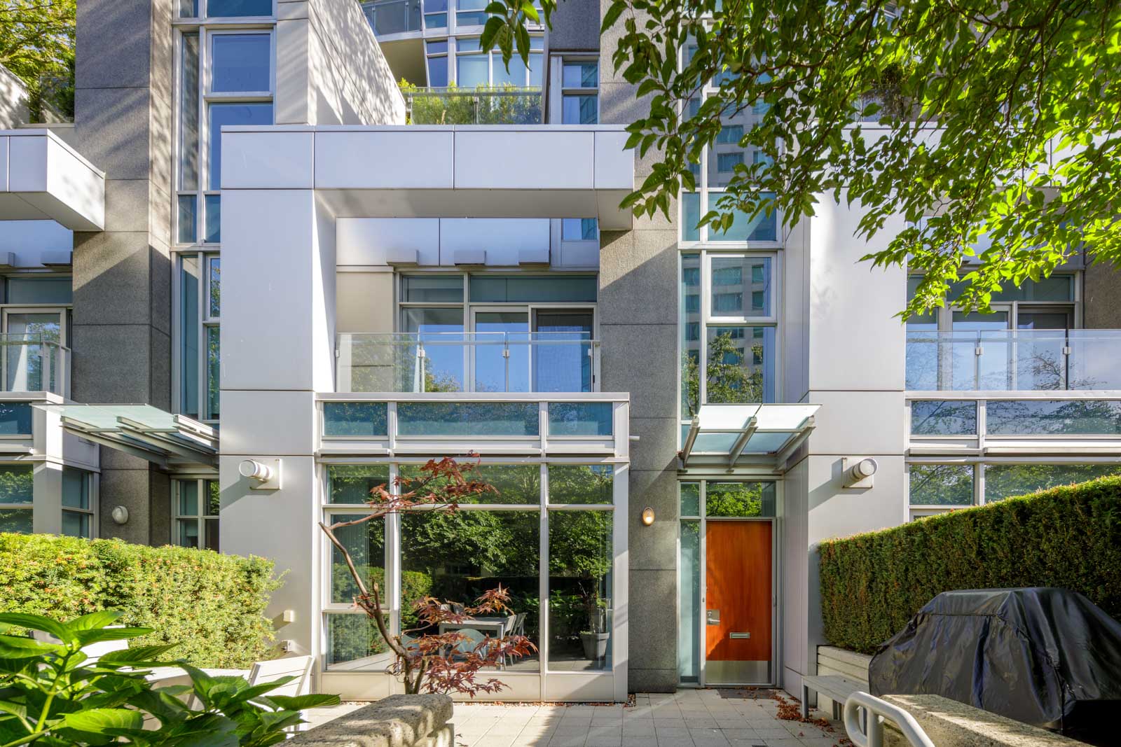 Modern apartment building with large windows, glass balconies, and a wooden entrance door, surrounded by greenery and outdoor seating area.