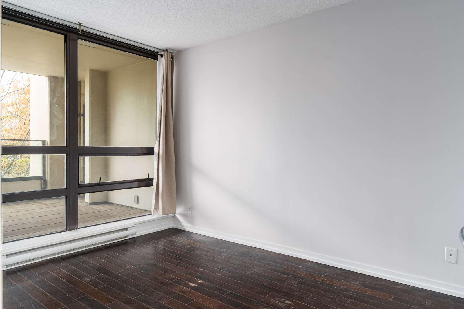 Empty room with dark wood floor, white walls, a large window, and a beige curtain partially open to a balcony.
