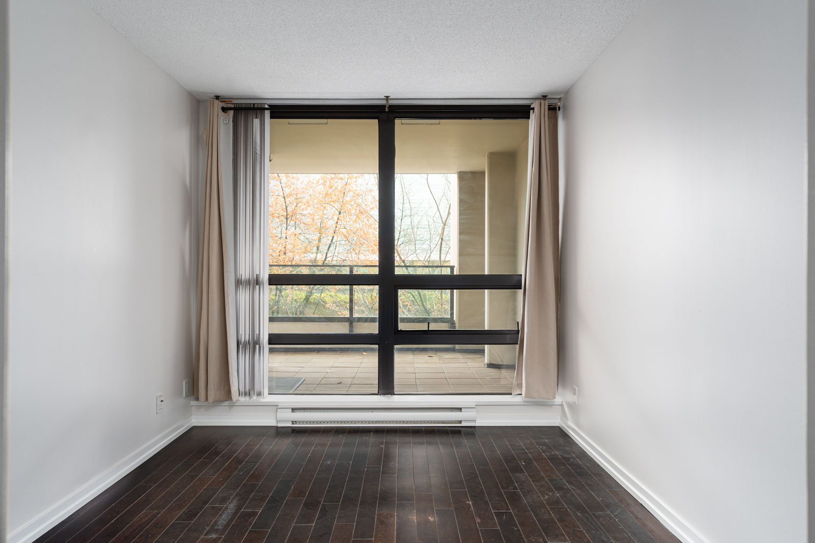 Empty room with dark wood flooring, white walls, and a large window with beige curtains overlooking a balcony and trees outside.