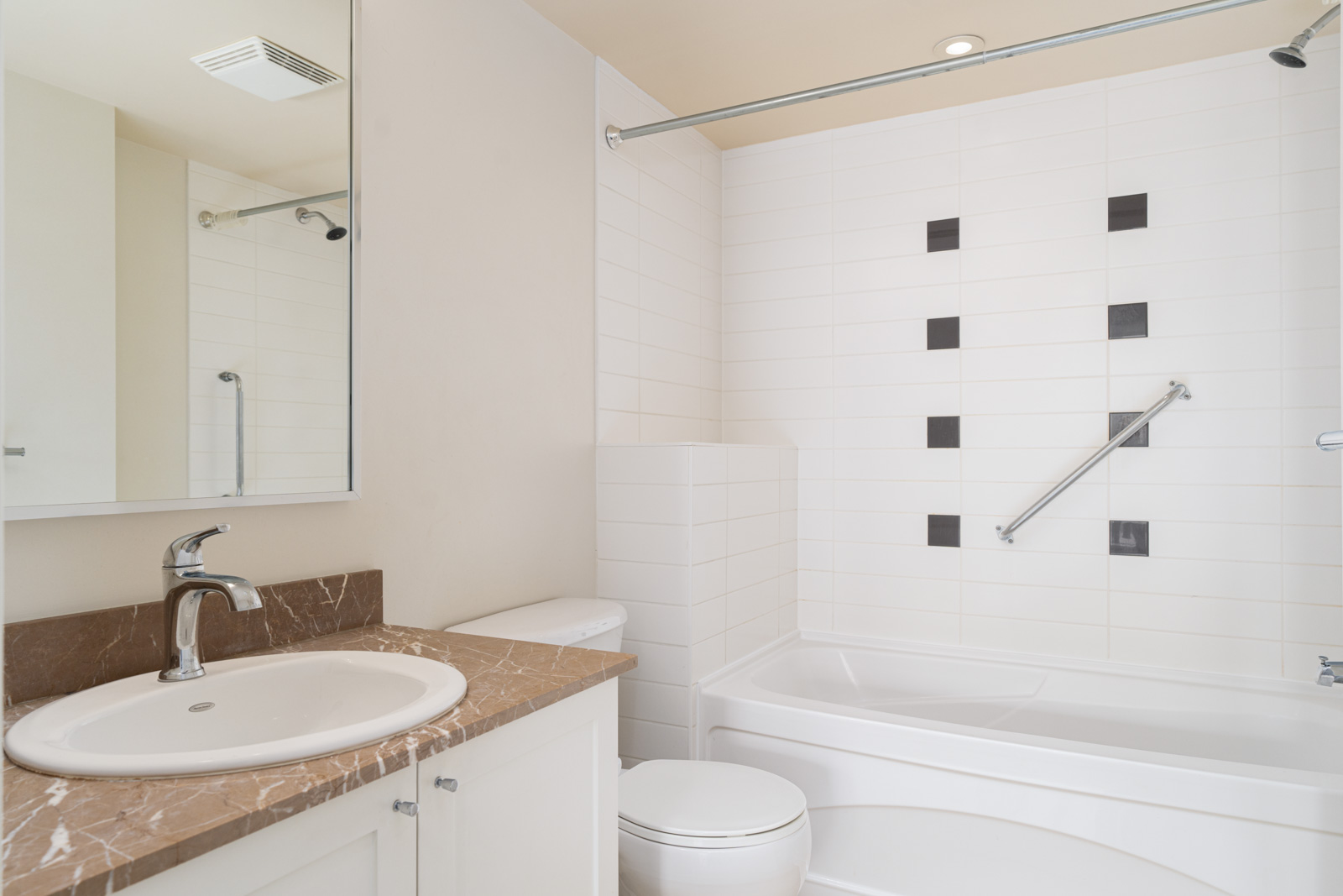 Modern bathroom with white tiles, a bathtub, a wall-mounted grab bar, a toilet, a brown marble countertop, and a sink with a mirror above.