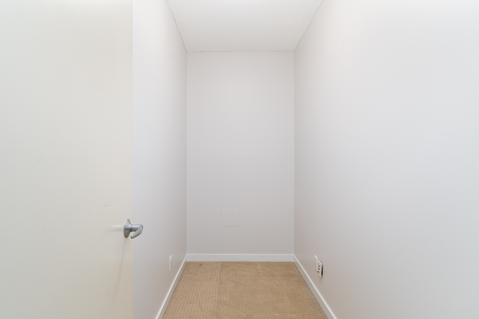 Small, empty, narrow room with plain white walls, beige carpet flooring, and a partially open door on the left side.