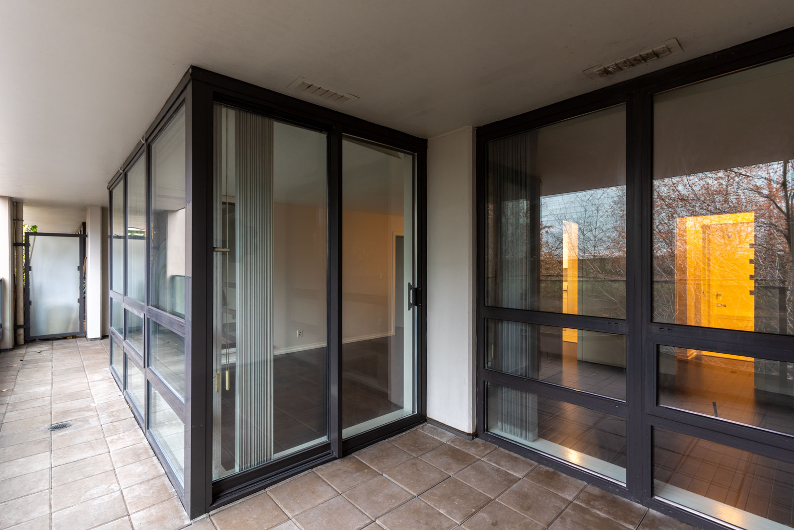 A modern glass-enclosed patio with large sliding doors, tiled floor, and an adjacent room visible through the windows.