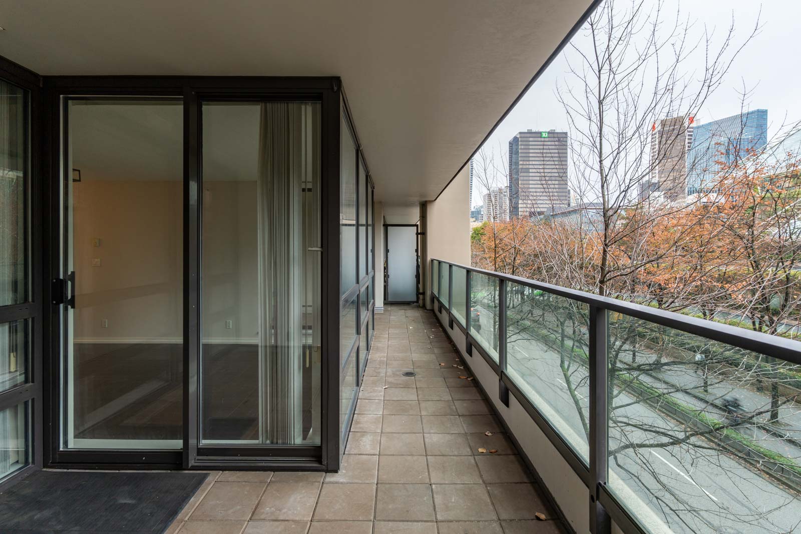A long balcony with glass railing, tiled floor, and sliding glass doors; bare trees and city buildings are visible in the background.