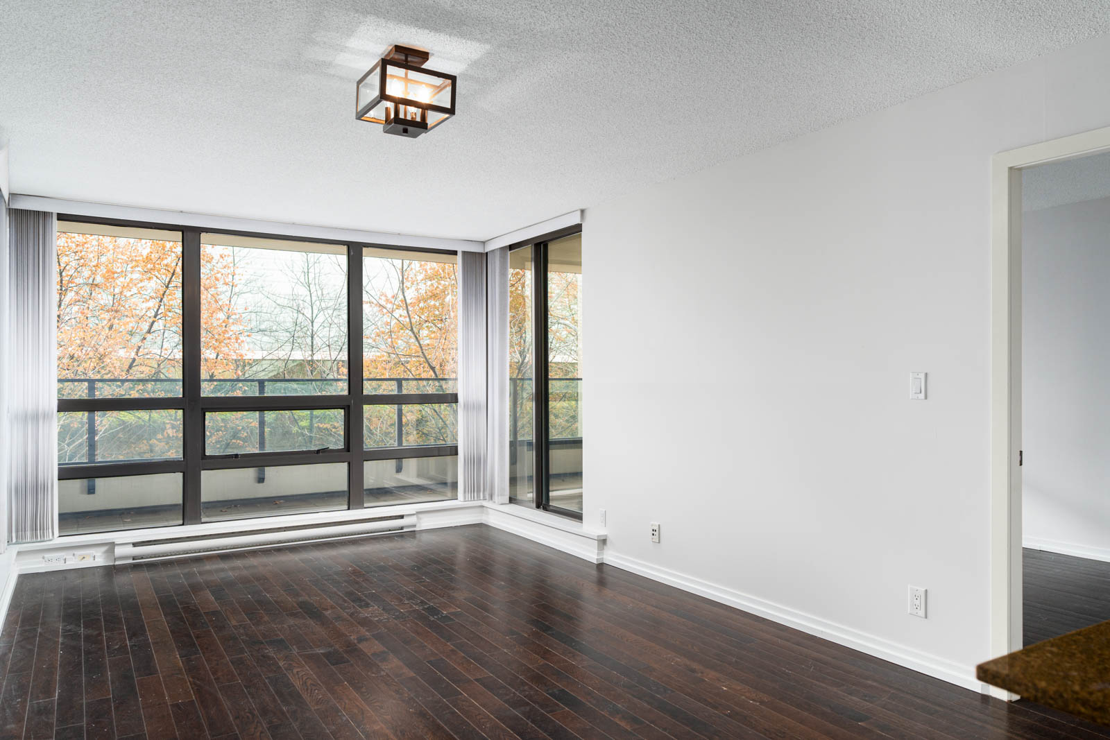 Empty living room with dark wood floors, large floor-to-ceiling windows, white walls, and a modern ceiling light fixture. Trees with autumn foliage are visible outside.