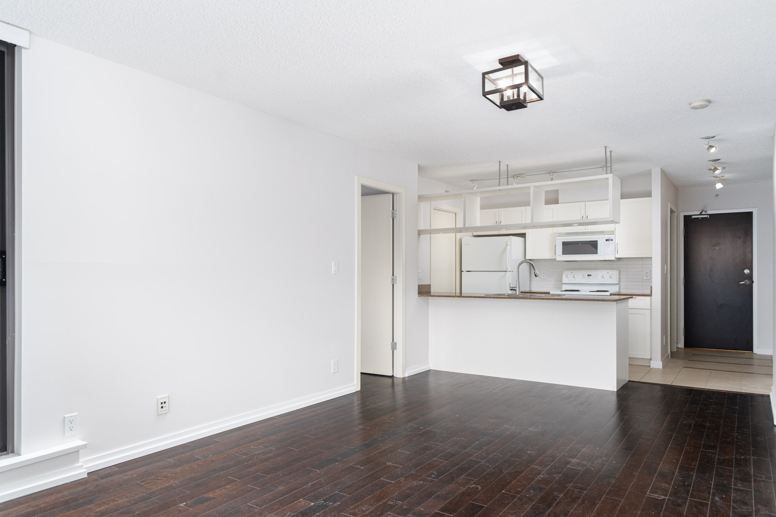 Unfurnished apartment with dark wood floors, white walls, and a kitchen featuring white appliances and cabinetry. A door leads to a hallway near the kitchen area.