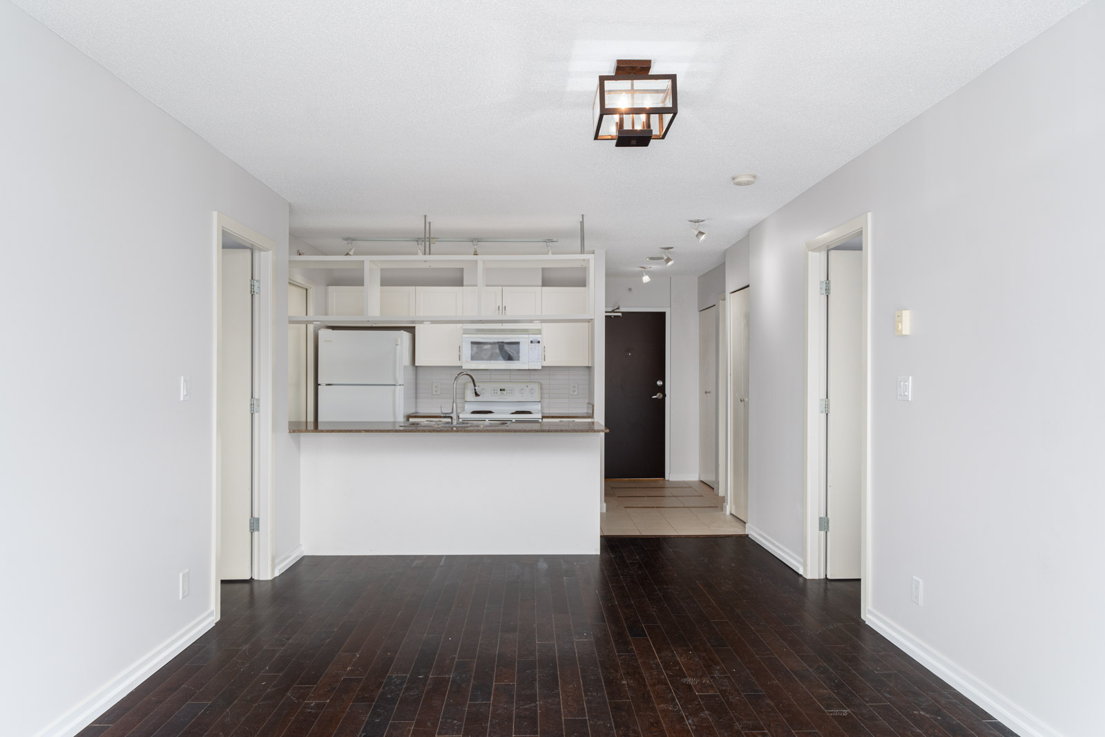 Unfurnished apartment with dark wood floors, white walls, an open kitchen with white cabinets, and two doorways on either side.