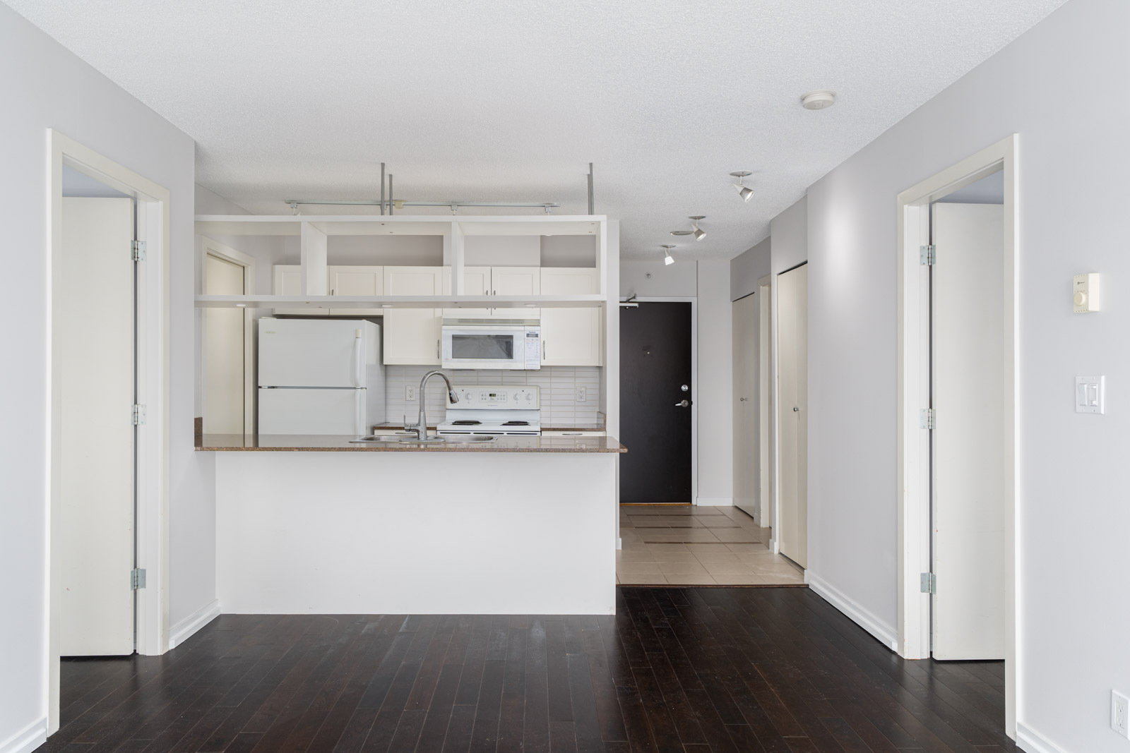 Empty modern kitchen with white cabinets, appliances, and a breakfast bar, viewed from a living area with dark wood flooring and light-colored walls.
