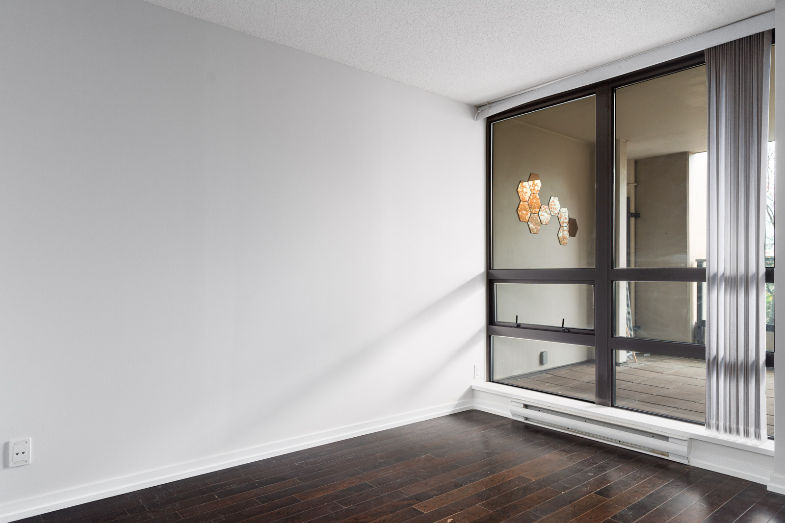 Empty room with white walls, dark wood flooring, and a large window with vertical blinds, reflecting a light fixture from outside.