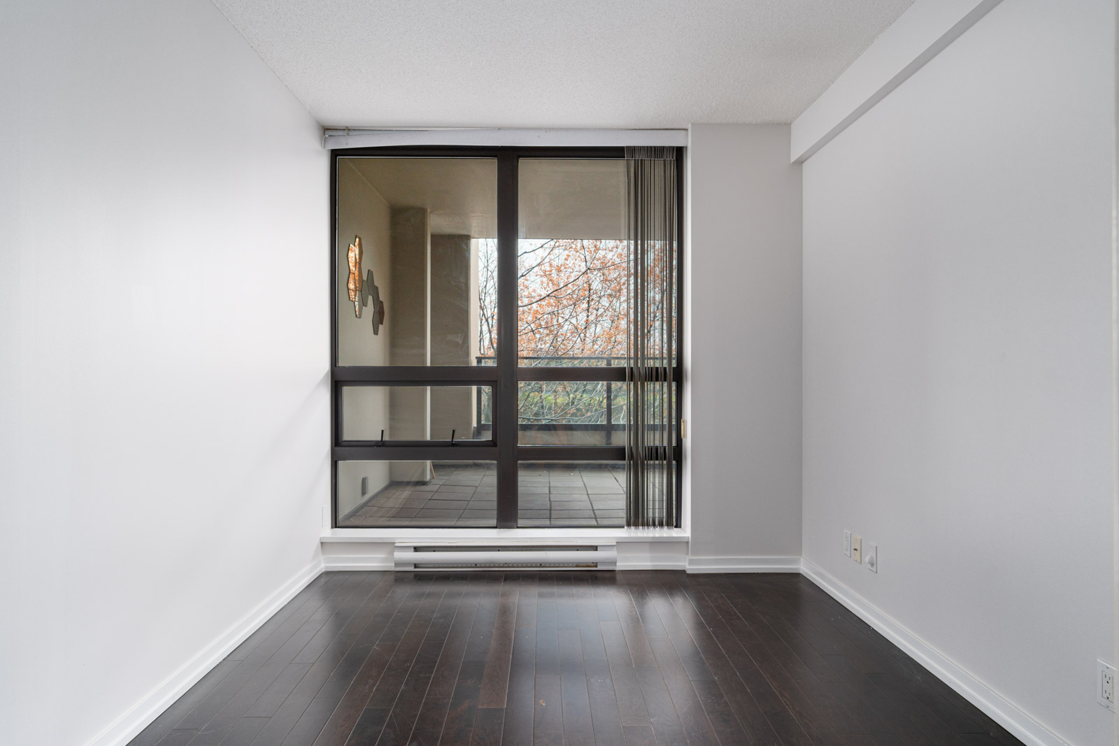 Empty room with white walls, dark wood flooring, and a large window with vertical blinds overlooking a balcony and trees outside.