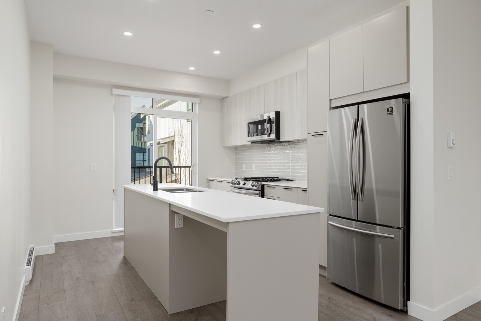 Modern kitchen with white cabinets, stainless steel appliances, an island with a sink, and wood flooring. Sliding glass door provides natural light.