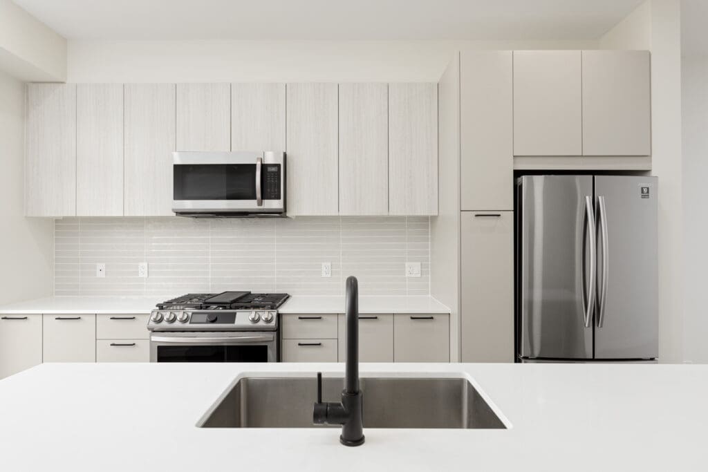 Modern kitchen with a stainless steel refrigerator, oven, and microwave, light cabinets, tile backsplash, and a black faucet over a white island sink.