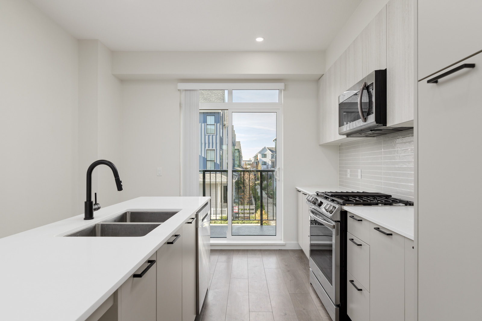 Modern kitchen with white cabinets, island sink, black faucet, stainless steel appliances, and large windowed door leading to a balcony with a view of neighboring buildings.