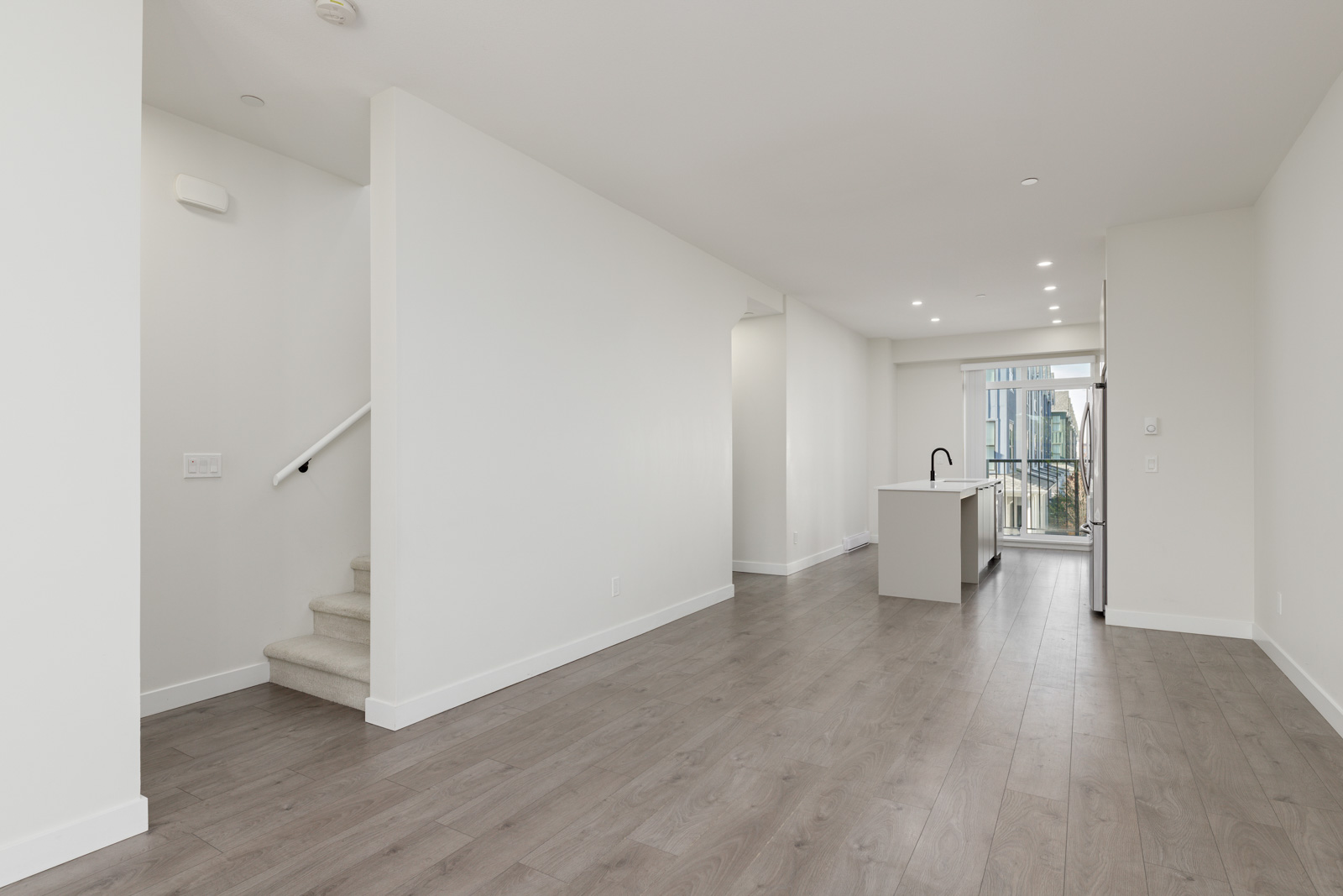 Unfurnished modern apartment with light-colored walls, wood flooring, a kitchen island, and a staircase leading upstairs. Large windows let in natural light.
