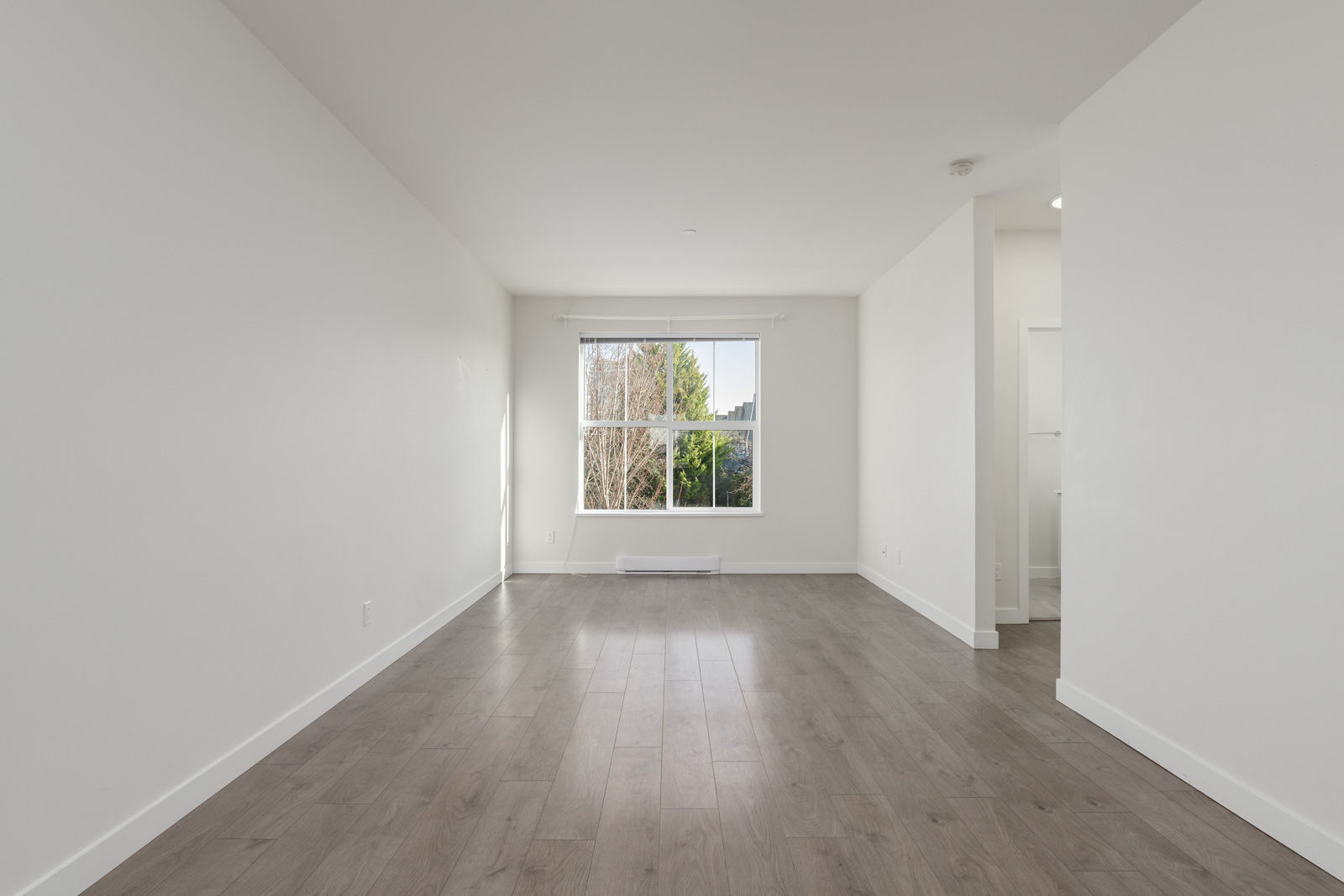 Unfurnished, empty room with white walls, wood flooring, a large window letting in natural light, and an open doorway leading to another area.