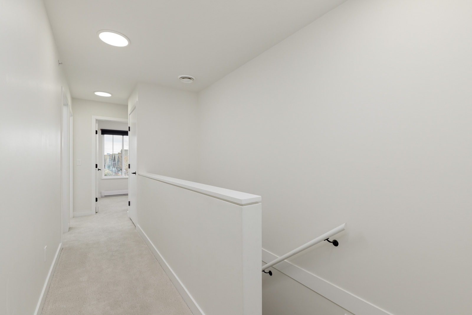 A bright, white hallway with carpeted floor, a railing on the right above a staircase, and a window at the far end letting in natural light.