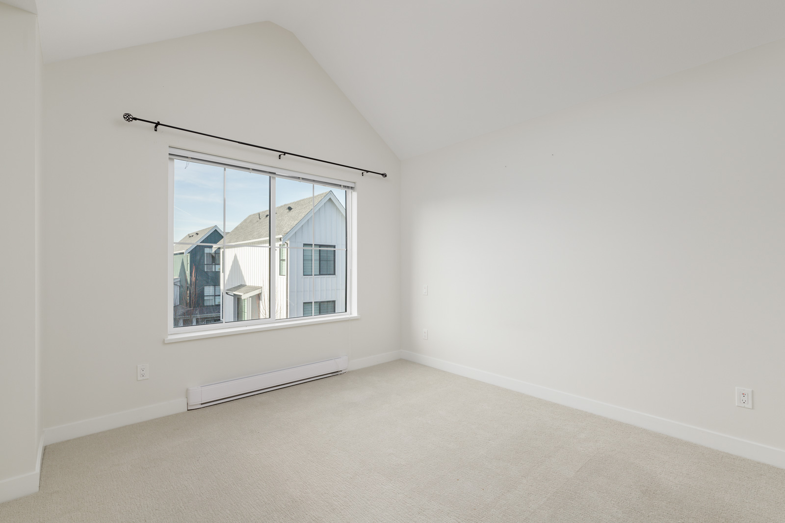 Empty room with beige carpet, white walls, a large window with a curtain rod, and a view of neighboring houses outside.