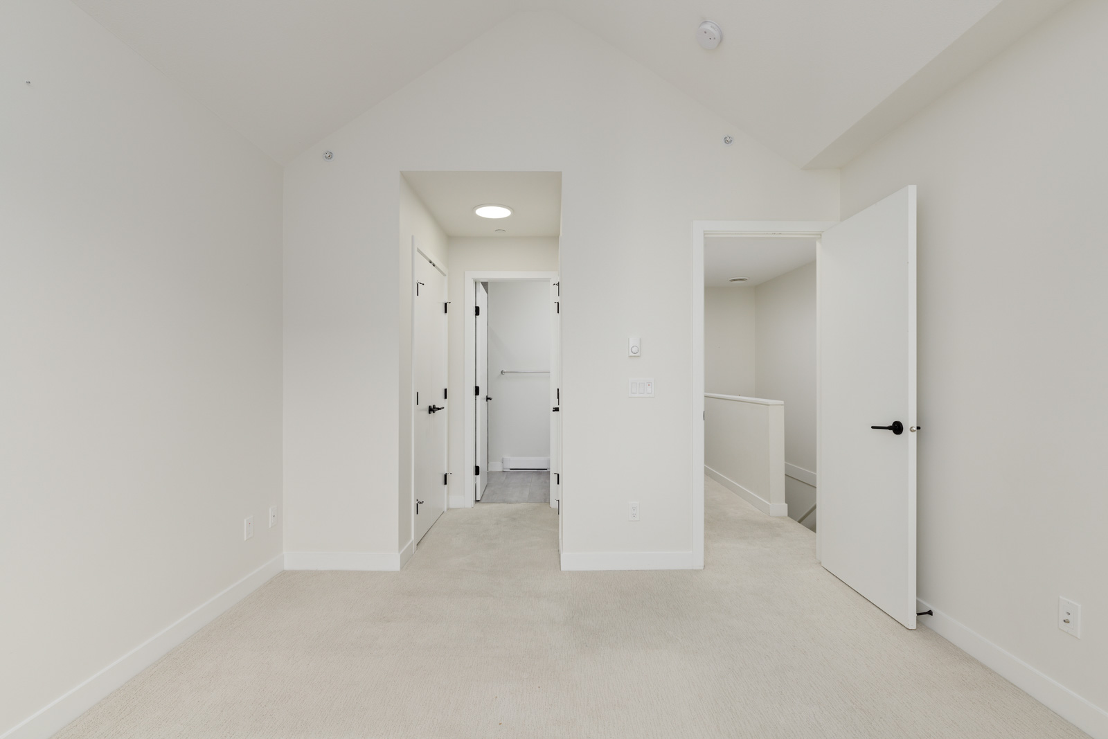 Empty room with light-colored carpet, white walls, high ceiling, and three open doors leading to adjoining areas and a hallway.