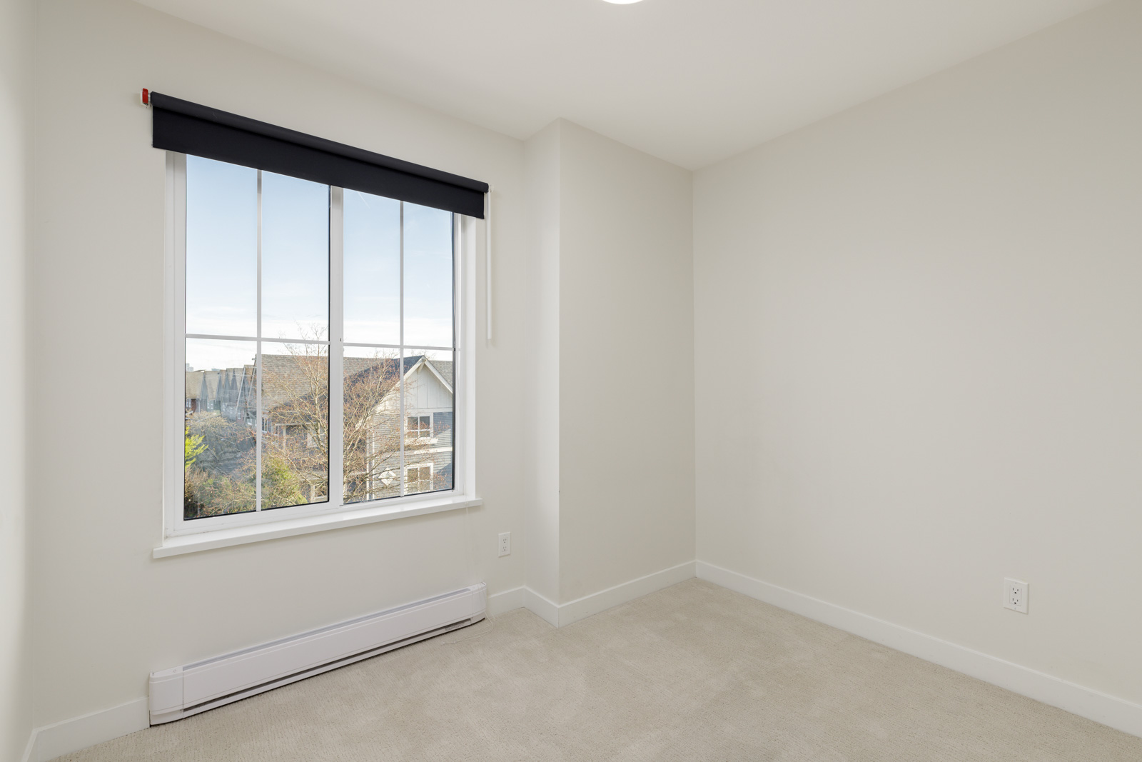 Empty room with light-colored walls and carpet, a large window with a black valance, and a view of houses and trees outside.