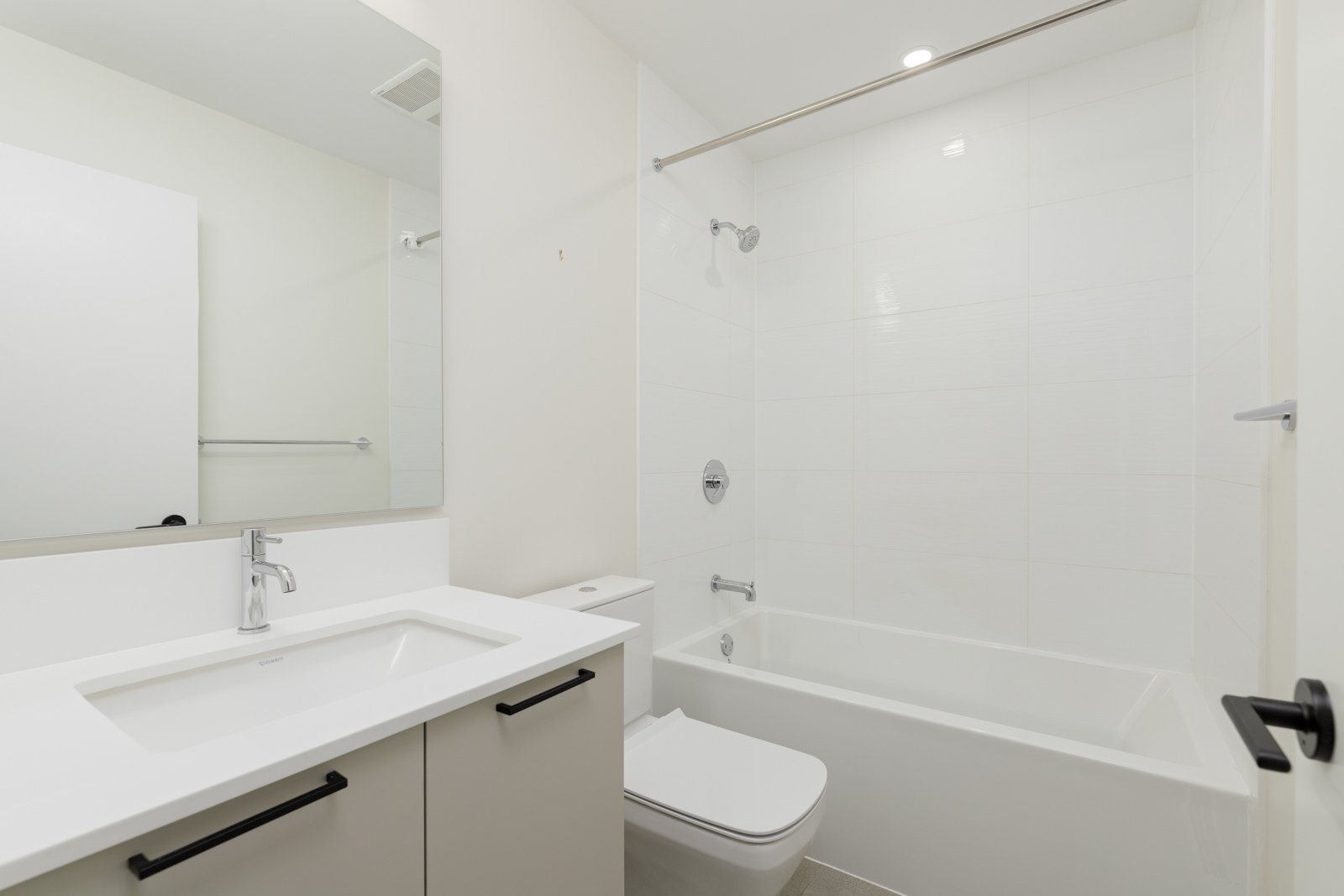 Modern bathroom with a white vanity, rectangular sink, wall mirror, toilet, and a bathtub with a showerhead and white tile surround.
