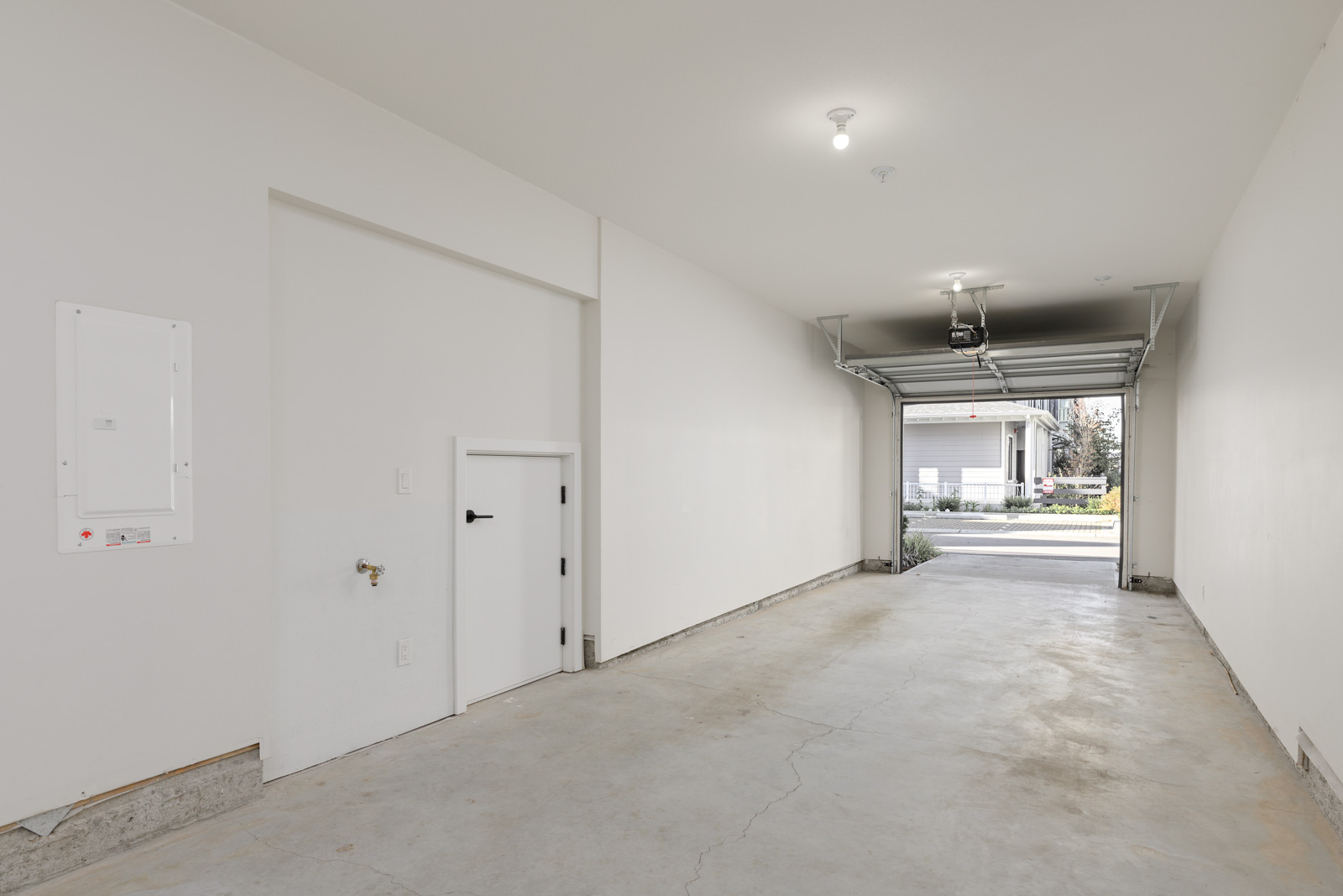 Empty single-car garage with a concrete floor, white walls, overhead lighting, a small utility door on the left, and an open garage door revealing a driveway and street outside.