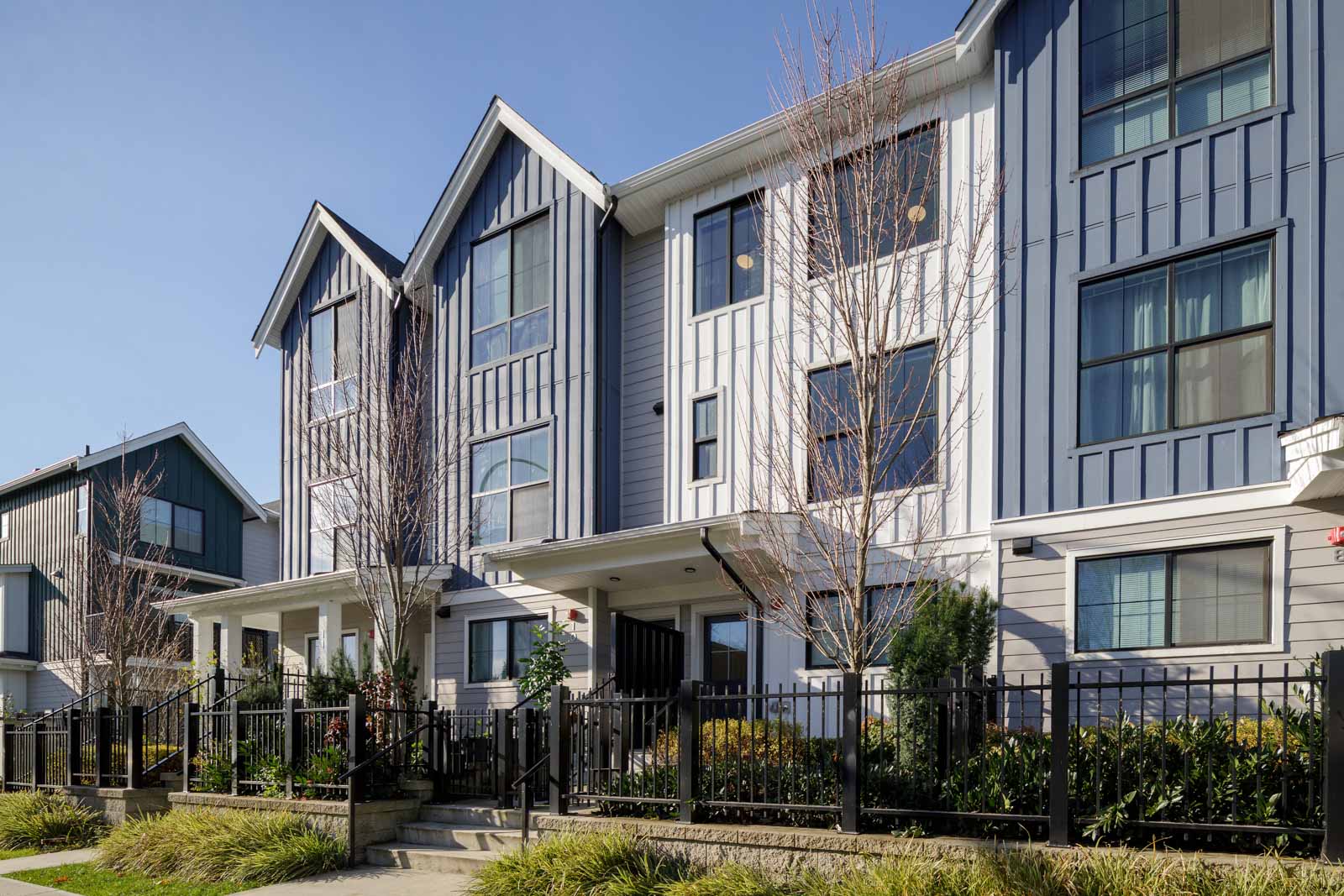 Modern three-story townhouse buildings with blue and white siding, large windows, small front gardens, and black metal fencing along the sidewalk.
