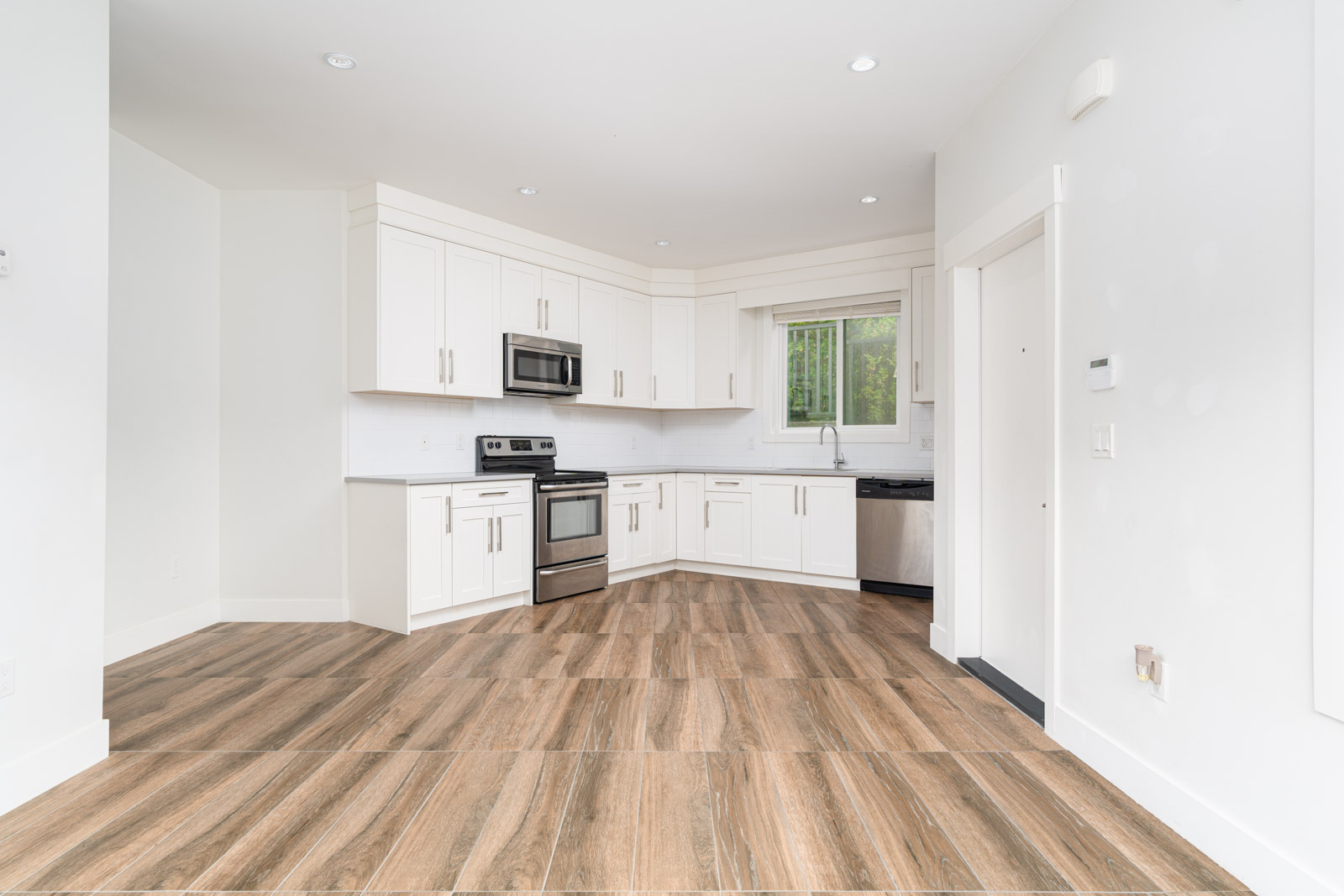 Modern kitchen with white cabinets, stainless steel appliances, and wood-look tile flooring, featuring a window above the sink and minimal decor.