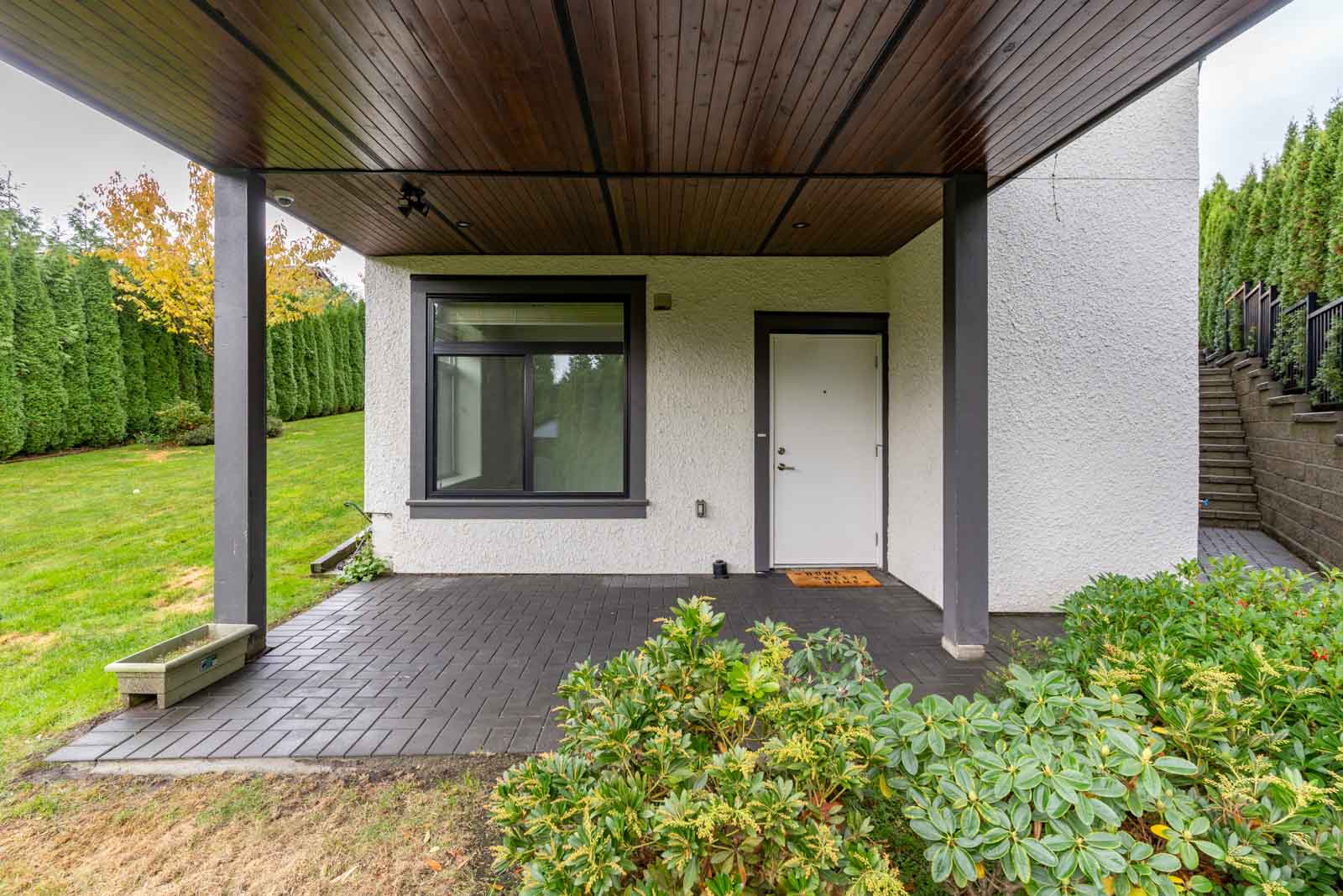 Covered patio area with a white exterior wall, black-trimmed window, white door, and tiled flooring, surrounded by greenery and a manicured lawn.